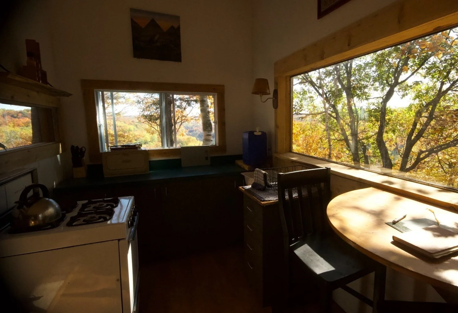 La cuisine sous le soleil bas d’automne : une cuisinière blanche au propane avec une bouilloire, un comptoir sombre le long du mur de fenêtres et une table en demi-lune sous une grande baie vitrée