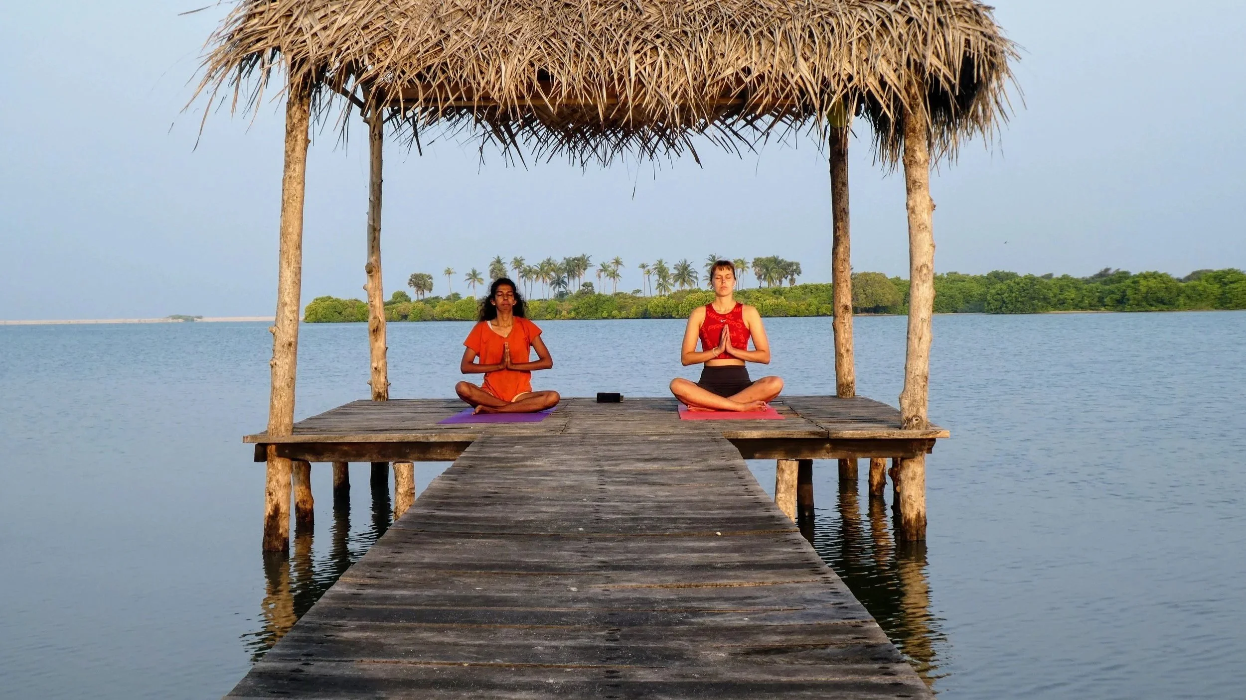 Two girls by the lagoon, practicing morning yoga on a wooden deck on the water