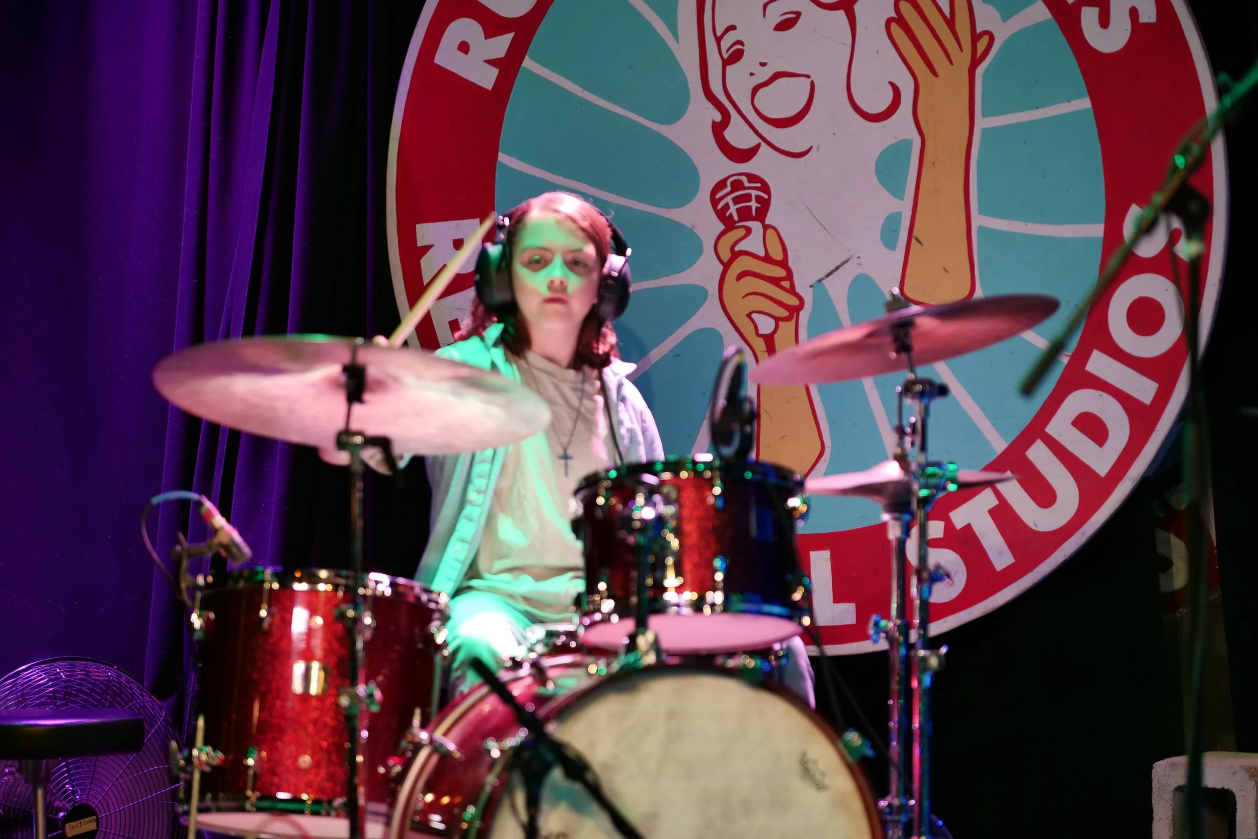 A young girl with headphones playing drums on stage, with a large colorful circular sign in the background featuring a woman holding a microphone and the text 'STUDIO'.
