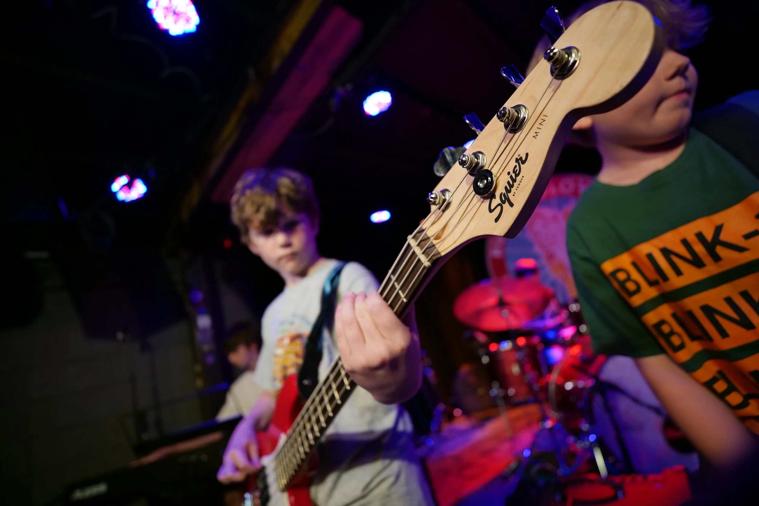 Young boys playing musical instruments on stage, with one boy prominently holding a guitar close to the camera, wearing a green BLINK-182 t-shirt, in a dimly lit venue with purple and blue stage lights.