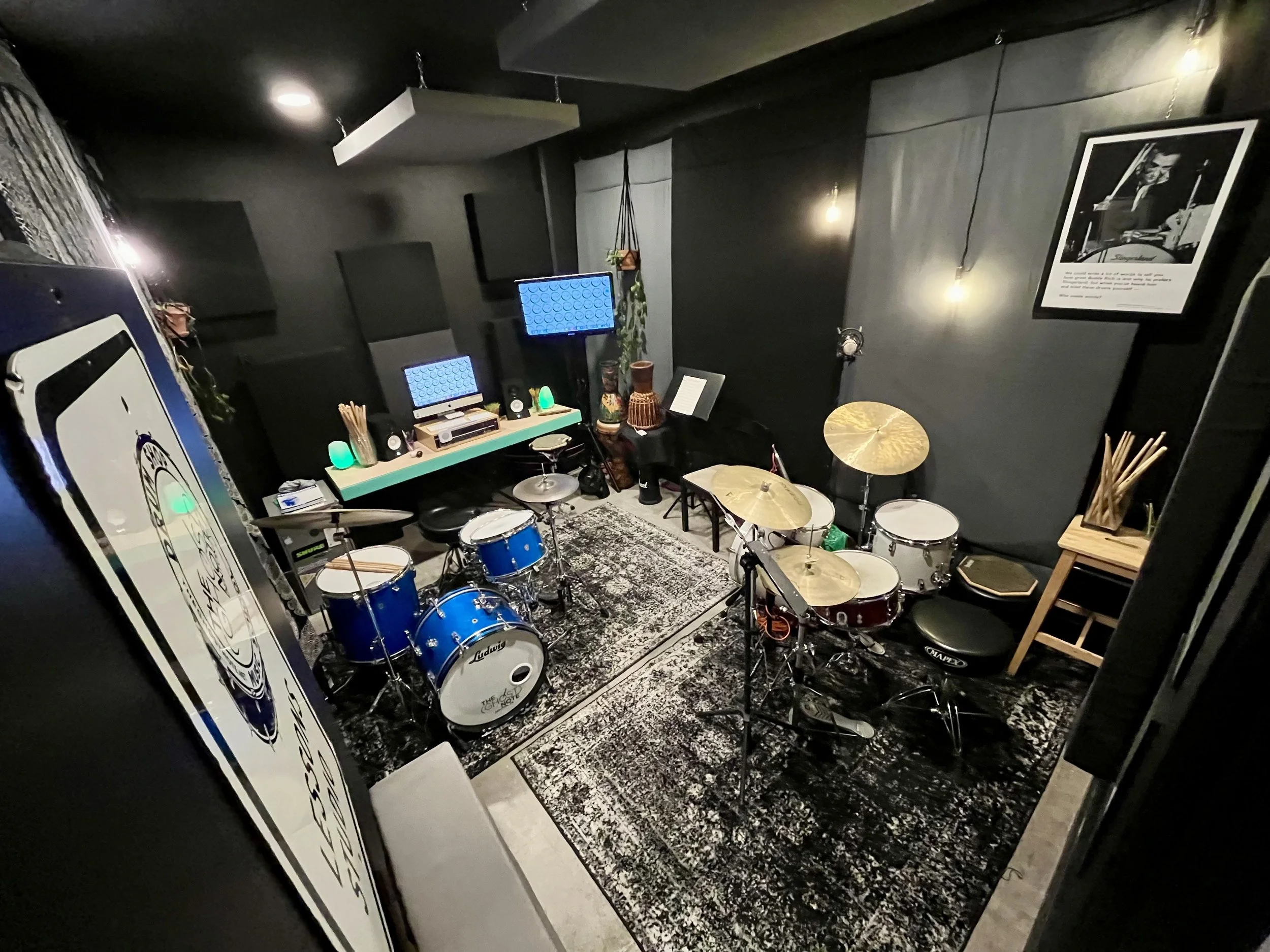 A music rehearsal room with drum kits, microphones, acoustic panels, and computer monitors, decorated with string lights, framed pictures, and eclectic decor.