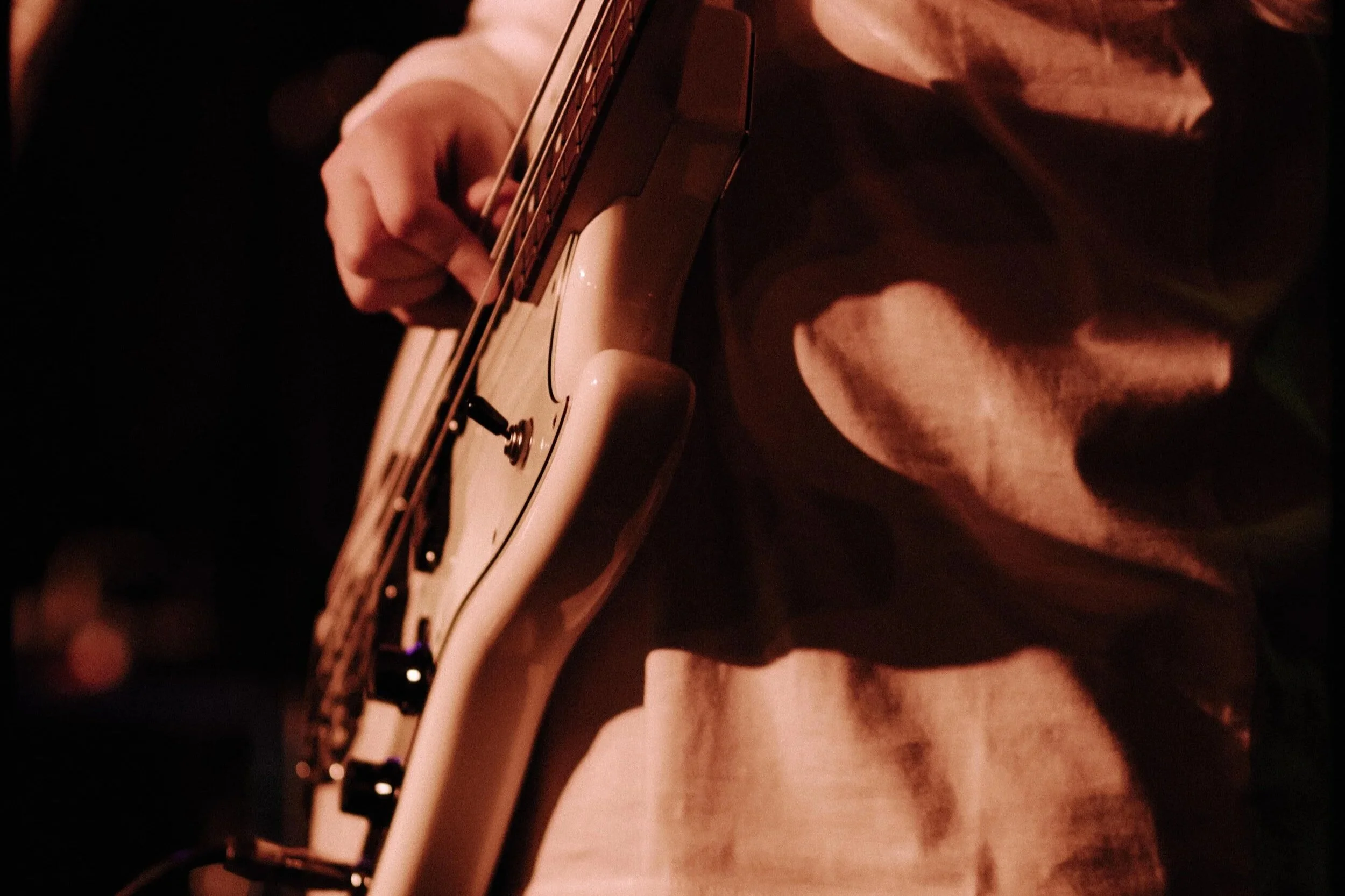 Close-up of a person playing an electric guitar in a dimly lit setting, focusing on their hand on the fretboard and part of the guitar body. Bass guitar lessons.