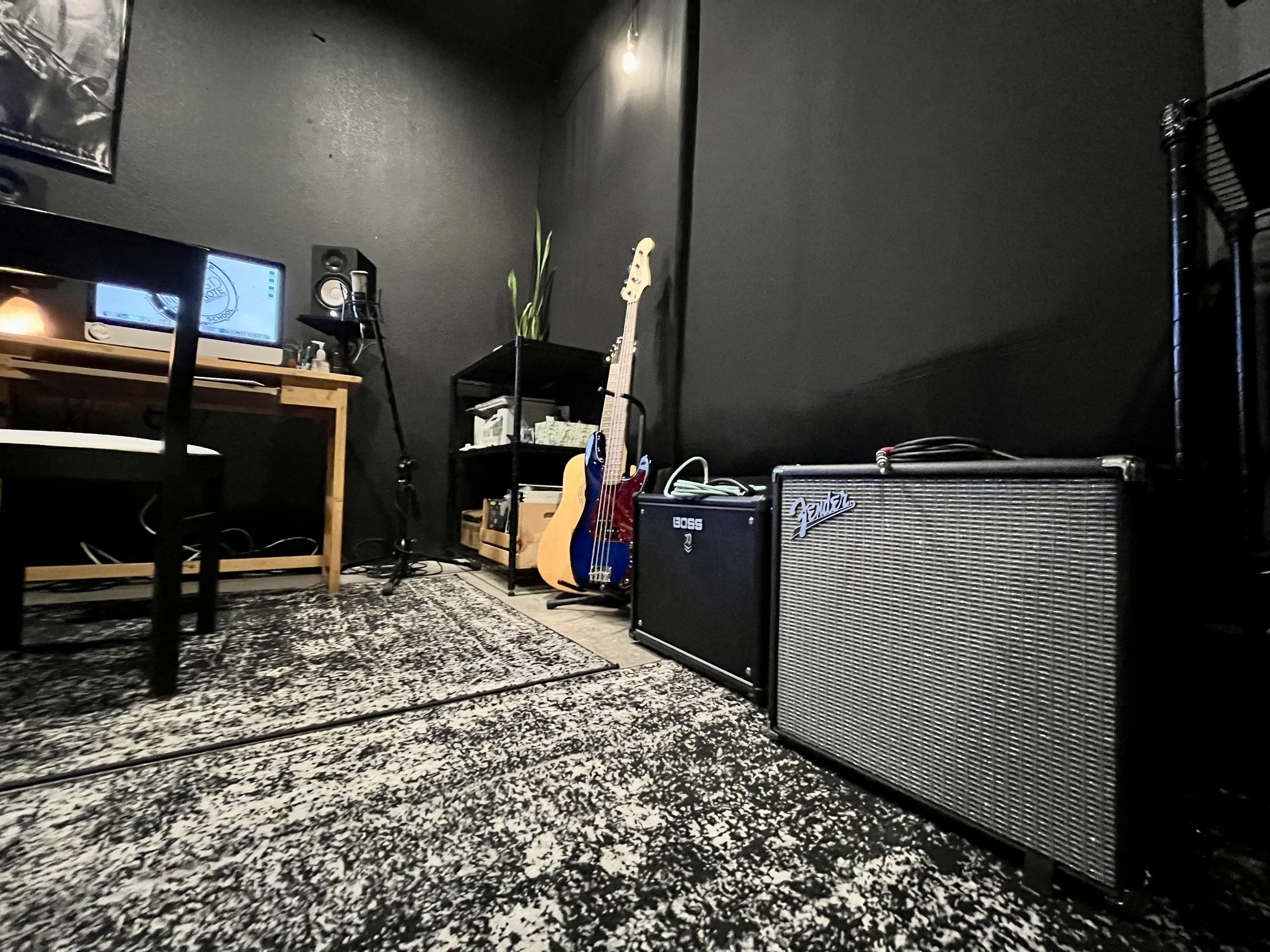 A room with a black wall, a wooden desk with a computer monitor, a speaker, and a chair. There are musical instruments including a bass guitar and an acoustic guitar. Two amplifiers are on the floor, one Fender and one Boss. A shelf with small items 