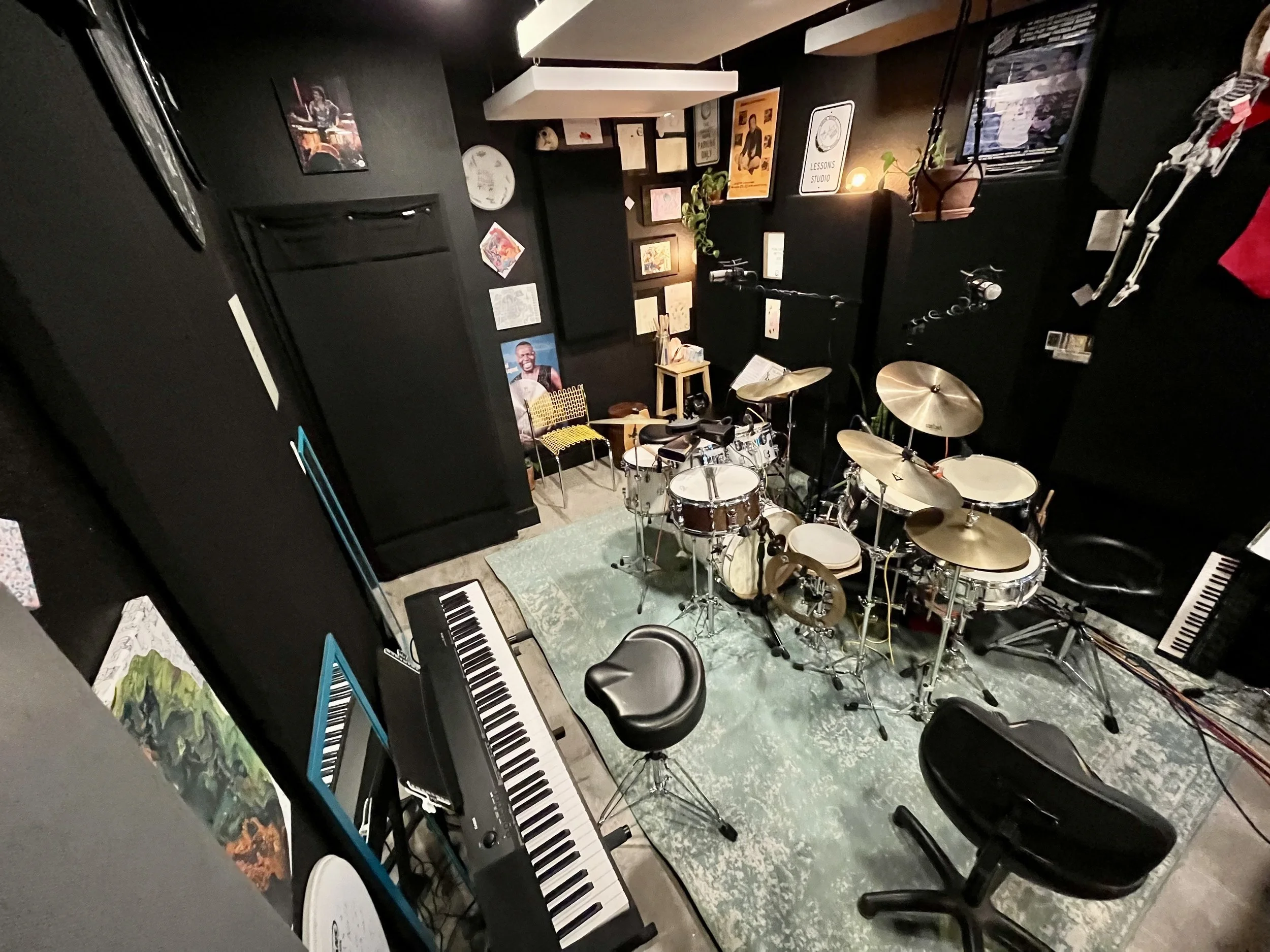 A music rehearsal room with a drum set, electronic keyboard, music stand, chairs, and various artwork and posters on black walls. The room has a vintage rug and a hanging potted plant.