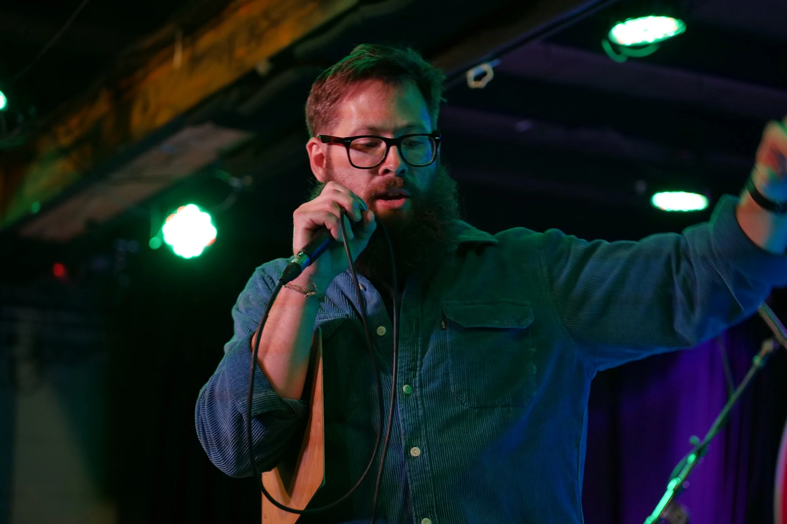 A man with glasses and a beard singing into a microphone on stage with green and purple lights.