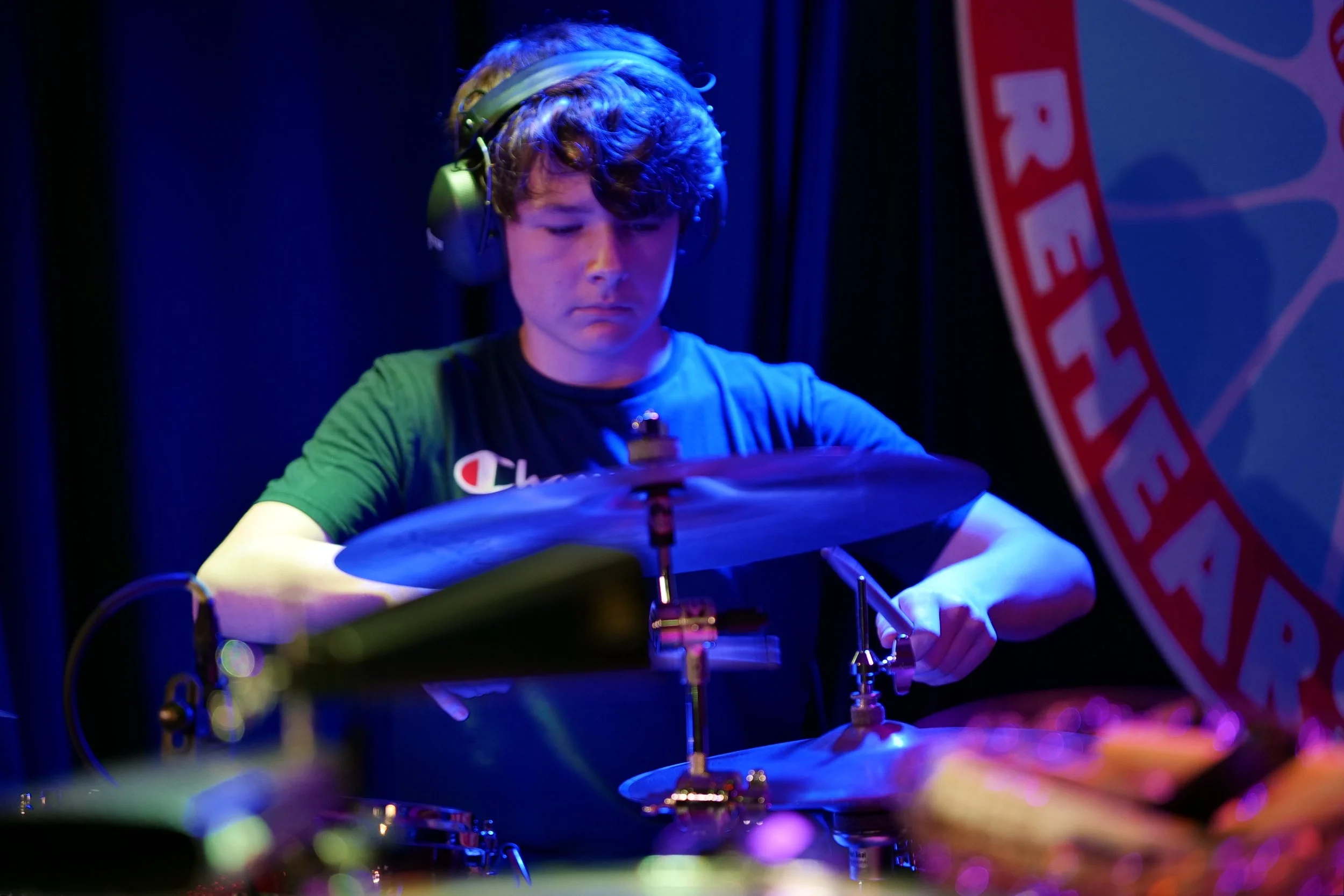 Young male musician wearing headphones playing drums on stage with a blue and purple lighting.