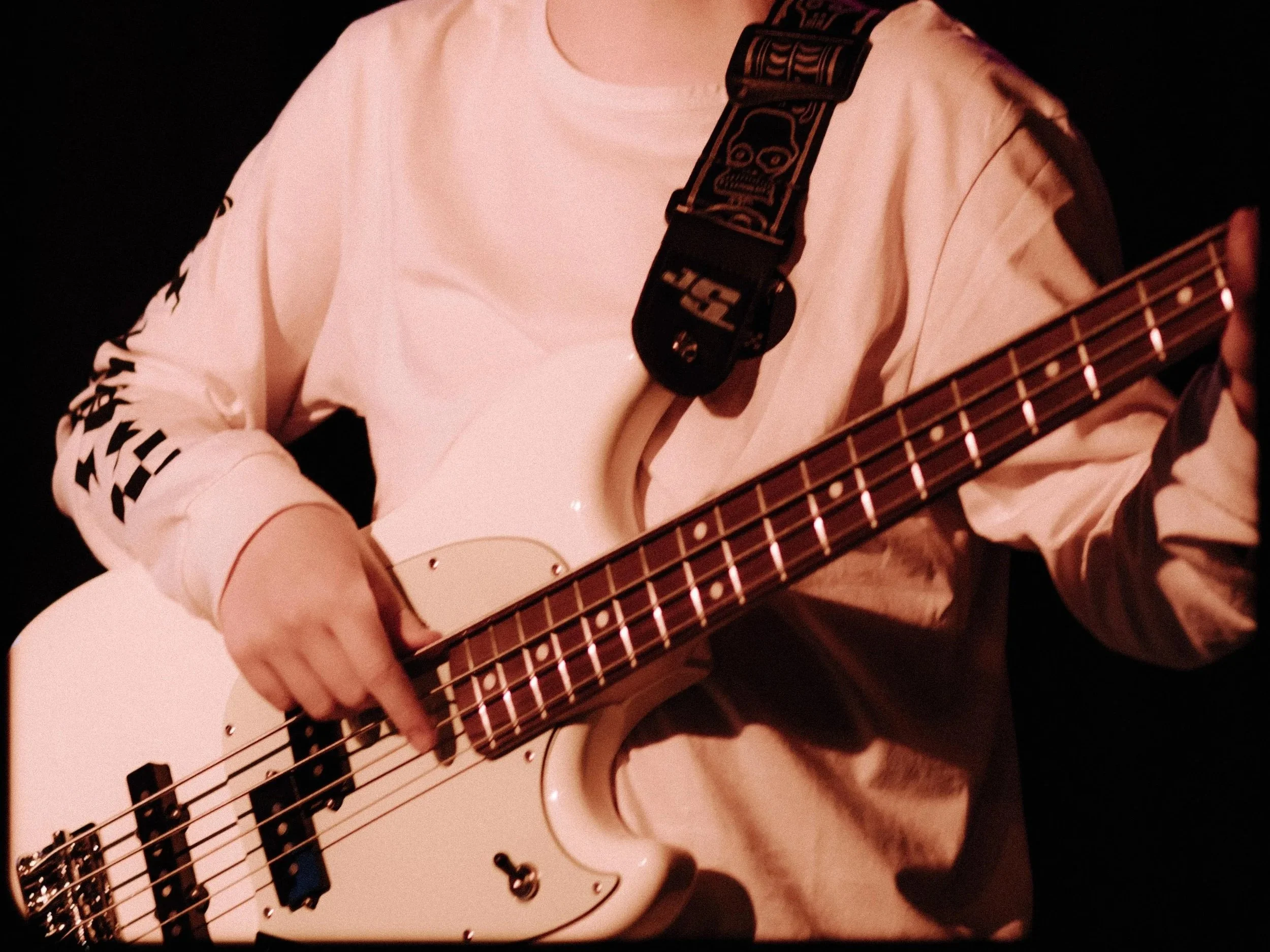 Person playing a white electric bass guitar, wearing a light-colored sweatshirt with text on the sleeves, a black strap with skull designs, in a dark setting. Bass guitar lessons. 