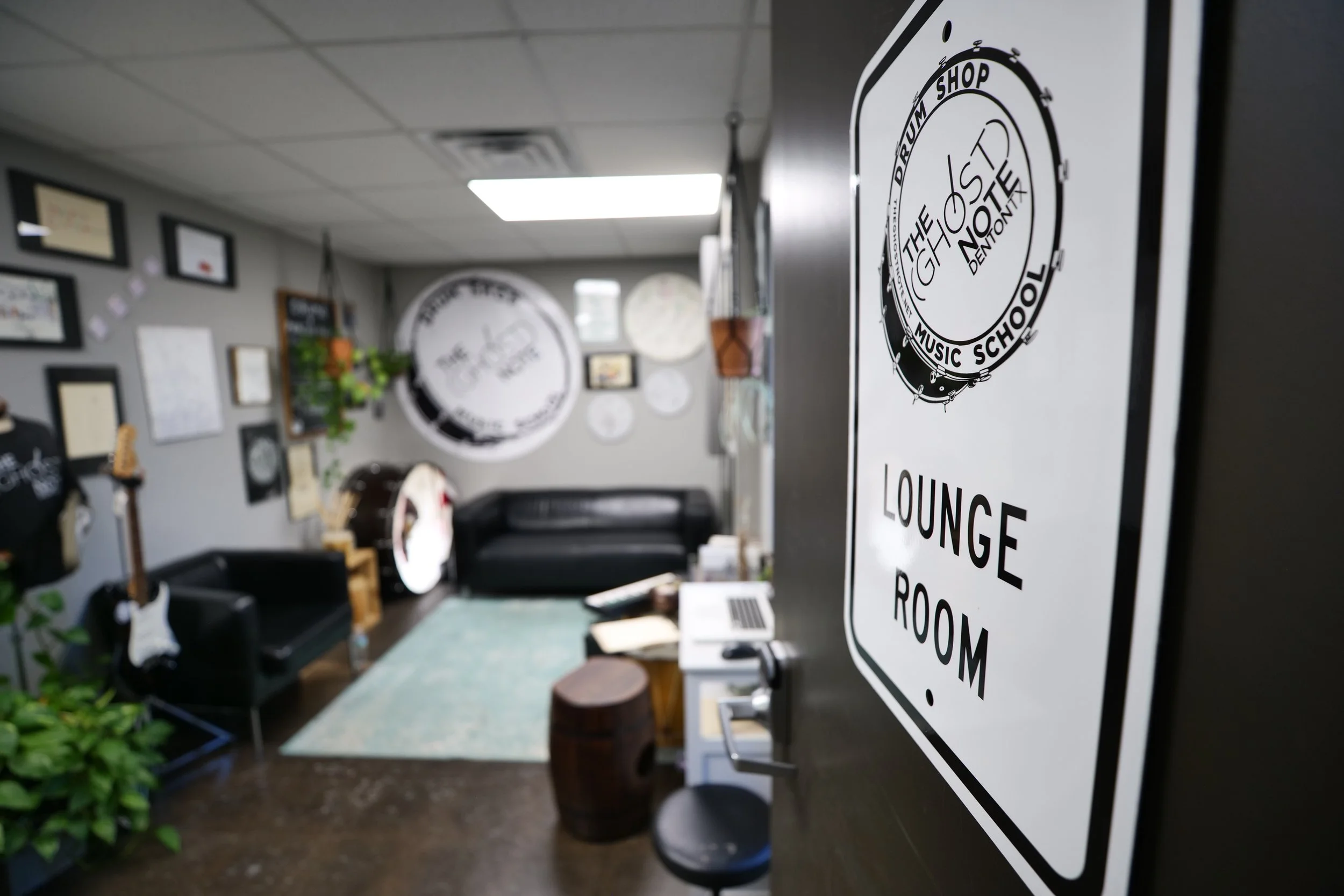 Interior of a music school lounge with a sign on a door that reads 'Lounge Room', featuring guitars, framed pictures, a couch, and a drum on the wall.