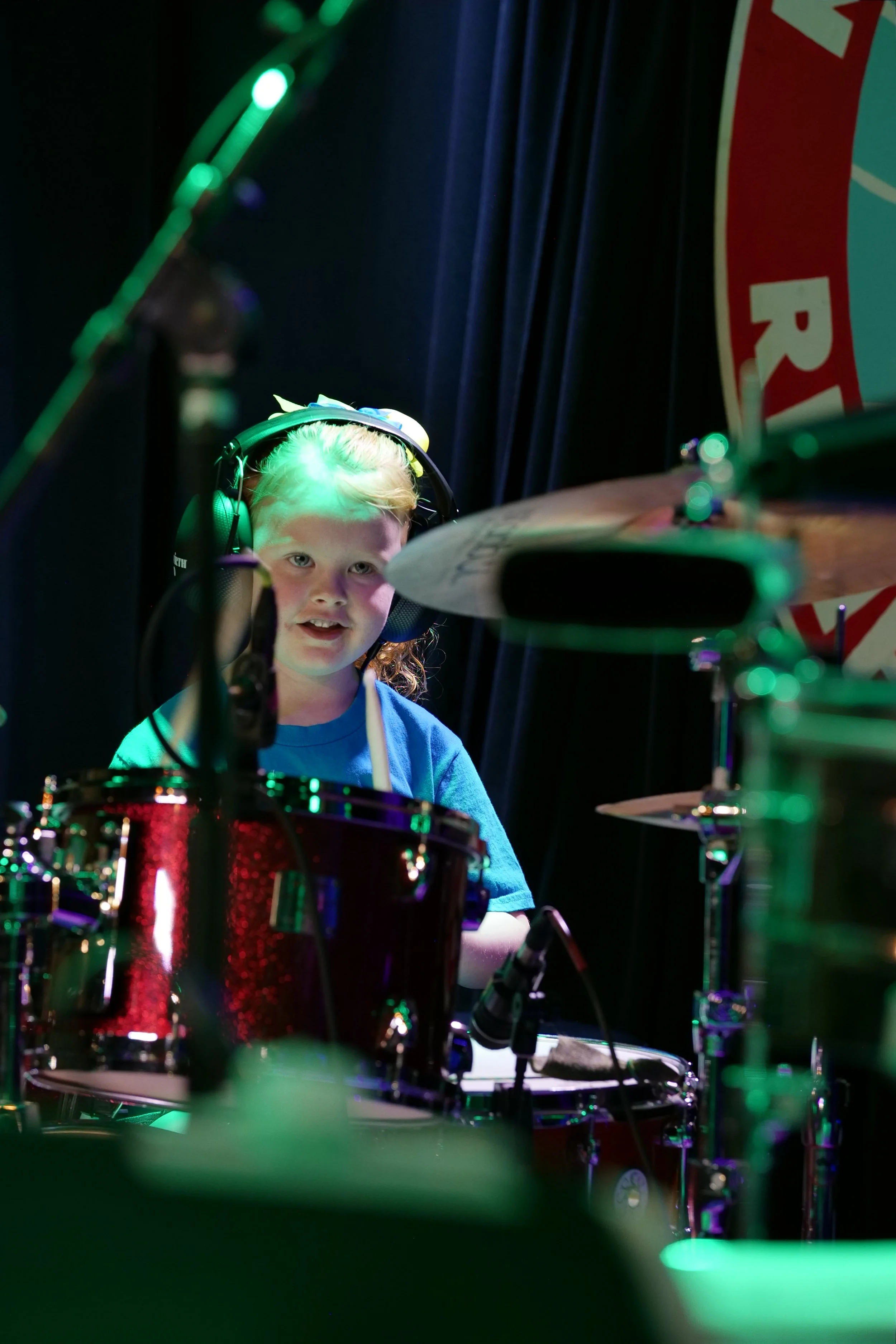 A young girl wearing headphones is sitting behind a drum set on a stage, looking at the camera.