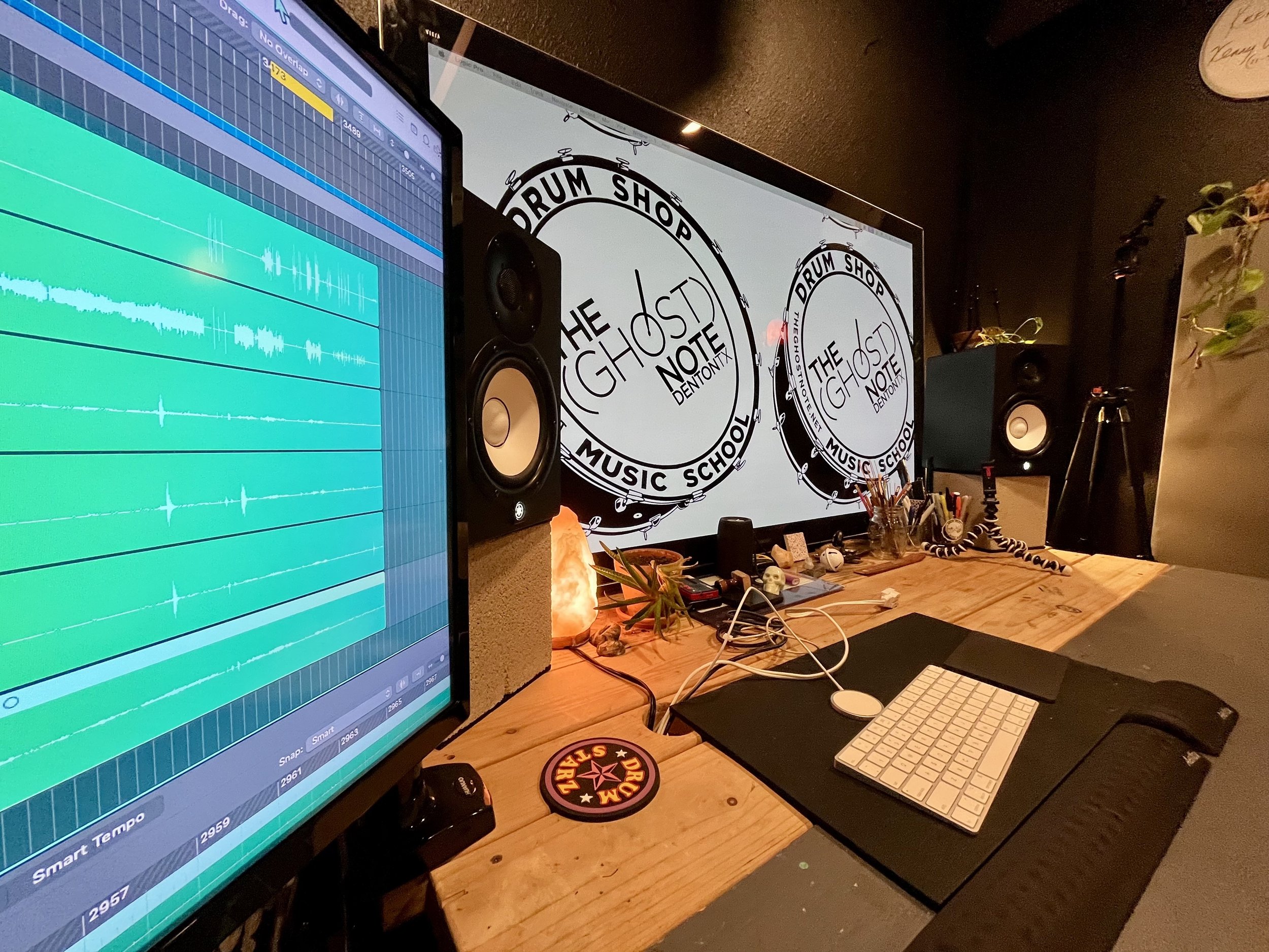 Music studio setup with a computer monitor displaying audio editing software, studio monitor speakers, a keyboard and mouse on a desk, and a large banner with the text 'The Ghost Note' and 'Drum Shop'.