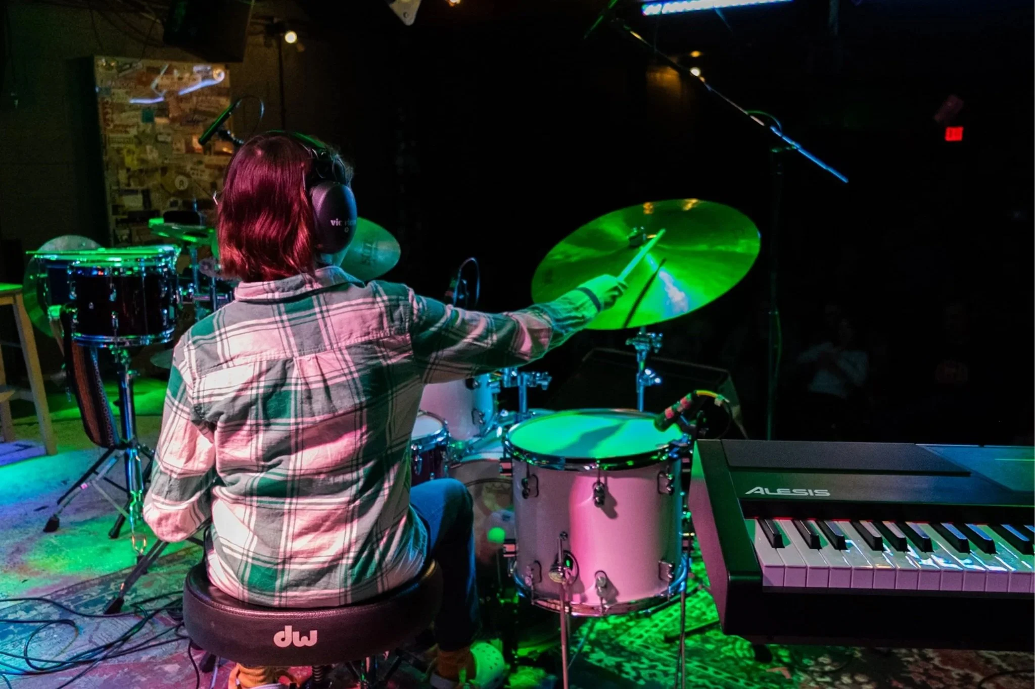 A person with shoulder-length hair wearing headphones and a plaid shirt is playing a drum set and keyboard on a stage with green and purple lighting.