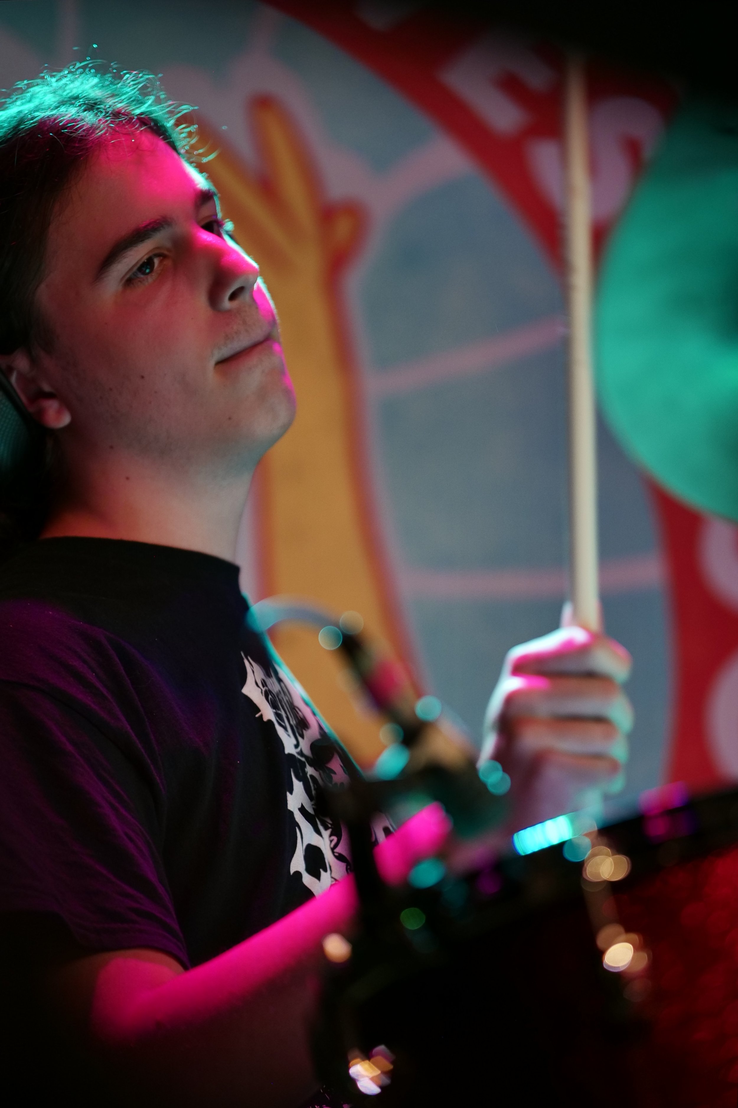 A young man plays drums in a colorful, vibrant setting, with pink and teal lighting, wearing a black T-shirt with a skull design, focused on his performance.