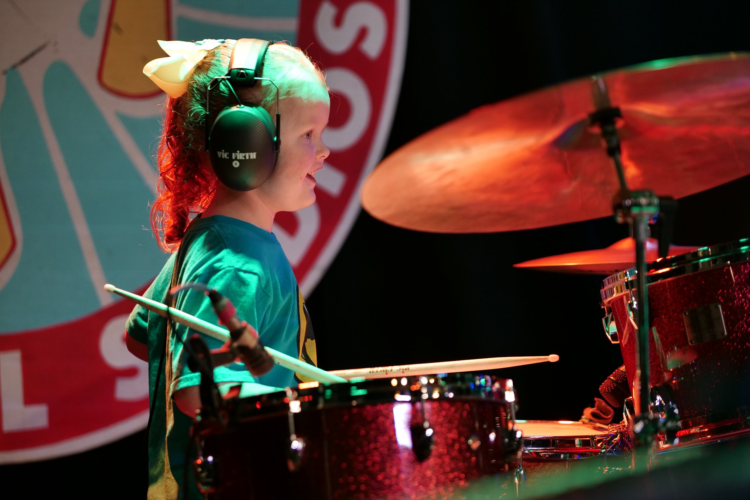 A young girl playing drums during a performance, wearing large black headphones and a blue shirt, with a colorful background.