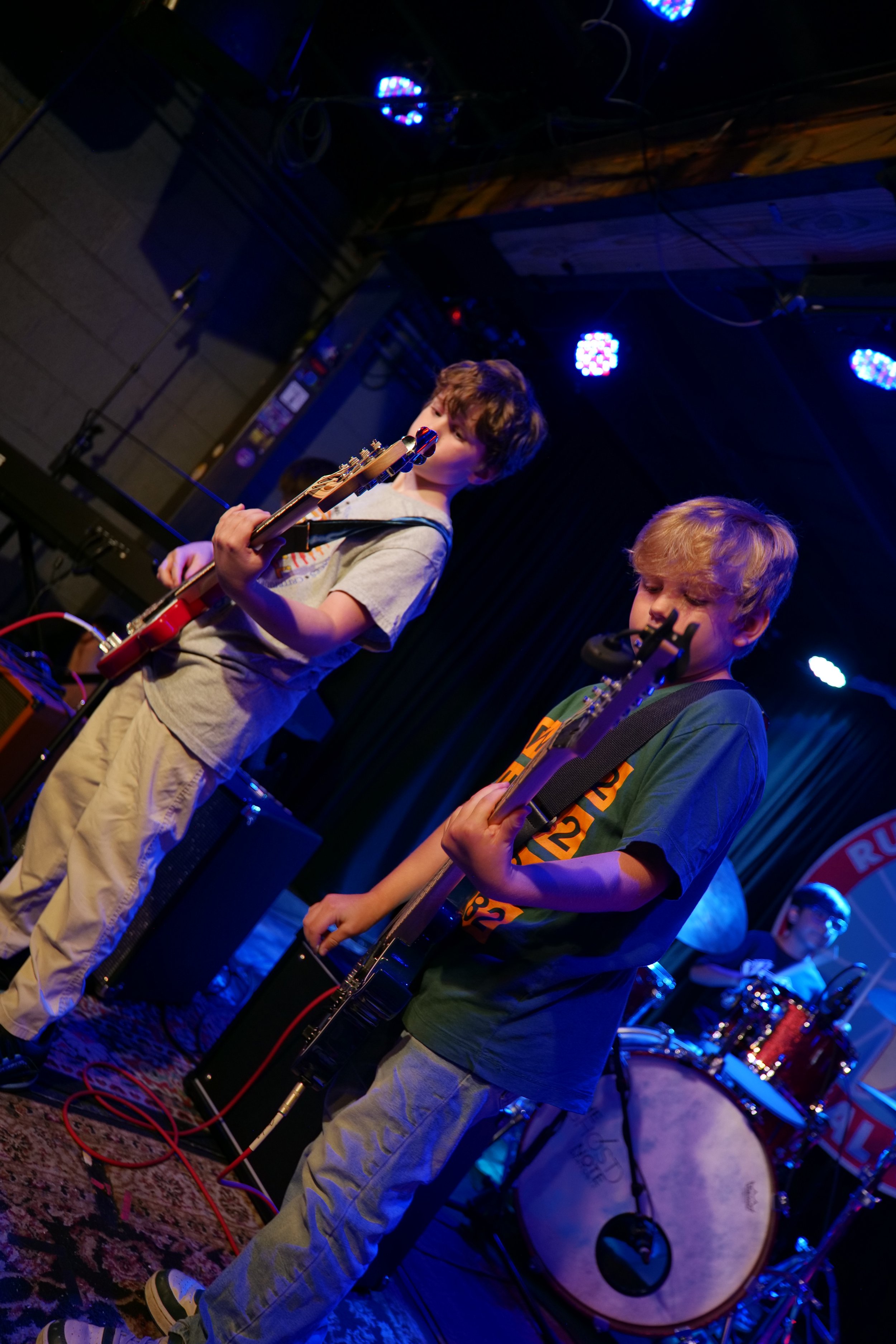 Two young boys playing electric guitars on stage with a drummer in the background, lit by colorful stage lights.