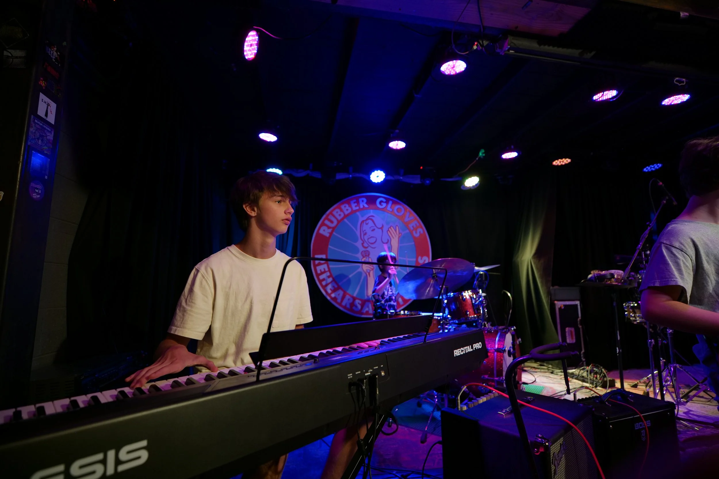 Young musician playing keyboard on stage with drummer in the background, illuminated by colorful stage lights, at a venue named Rubber Gloves. Student performance. 