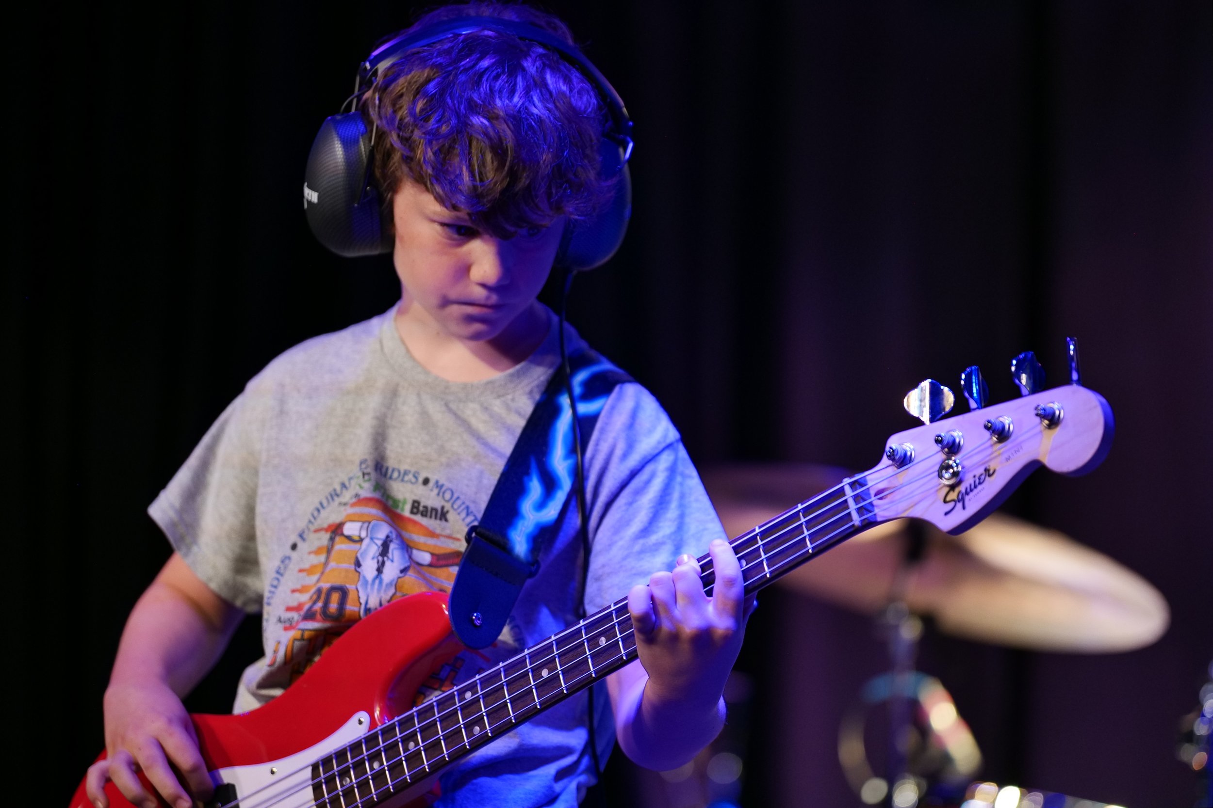 Young boy wearing headphones playing a red bass guitar on stage with a drum set in the background.