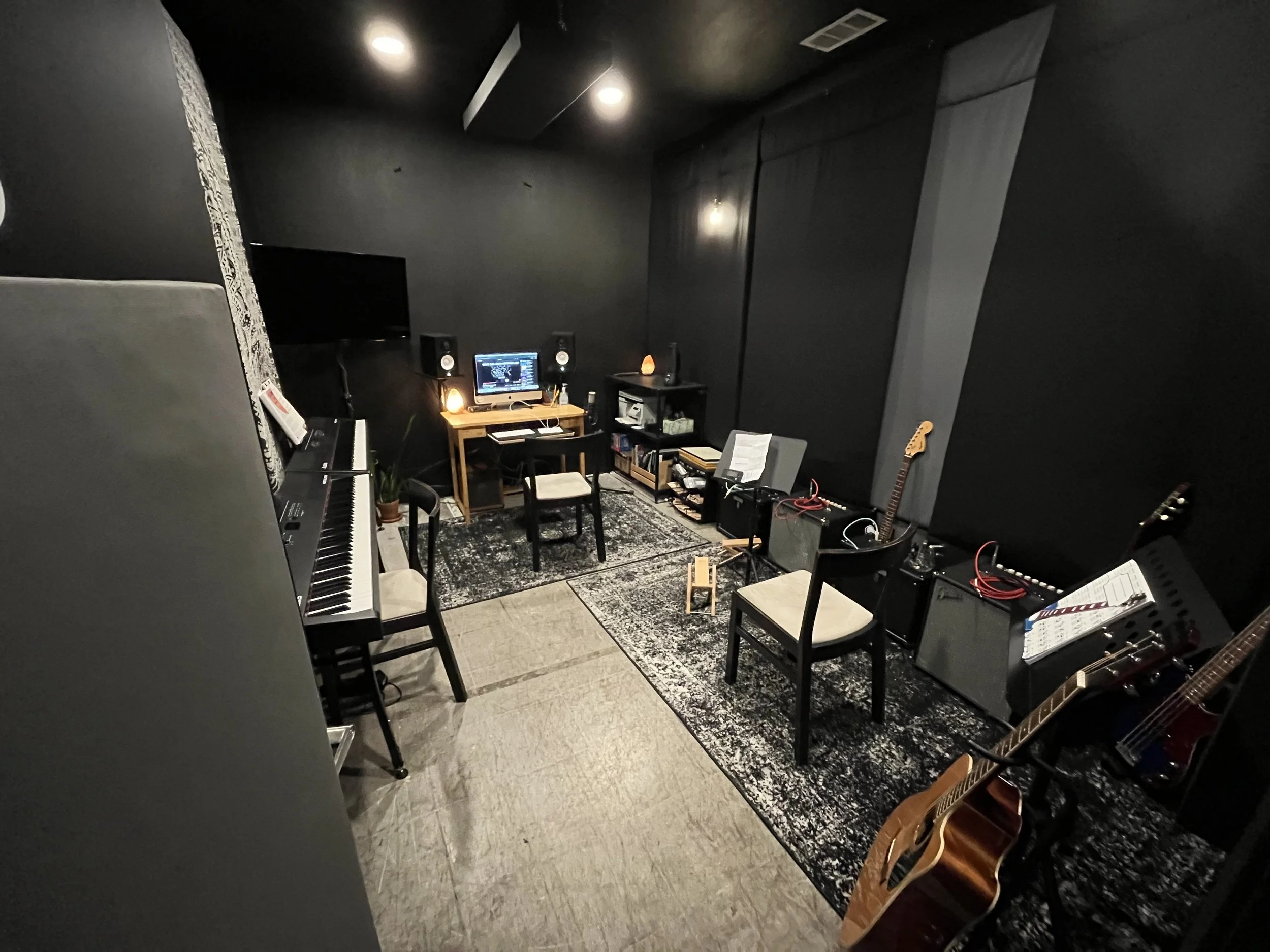 Music studio with black walls, a digital keyboard, two guitars, a computer on a small desk, two chairs, speakers, music equipment, and a TV. Music lesson studio Denton, TX