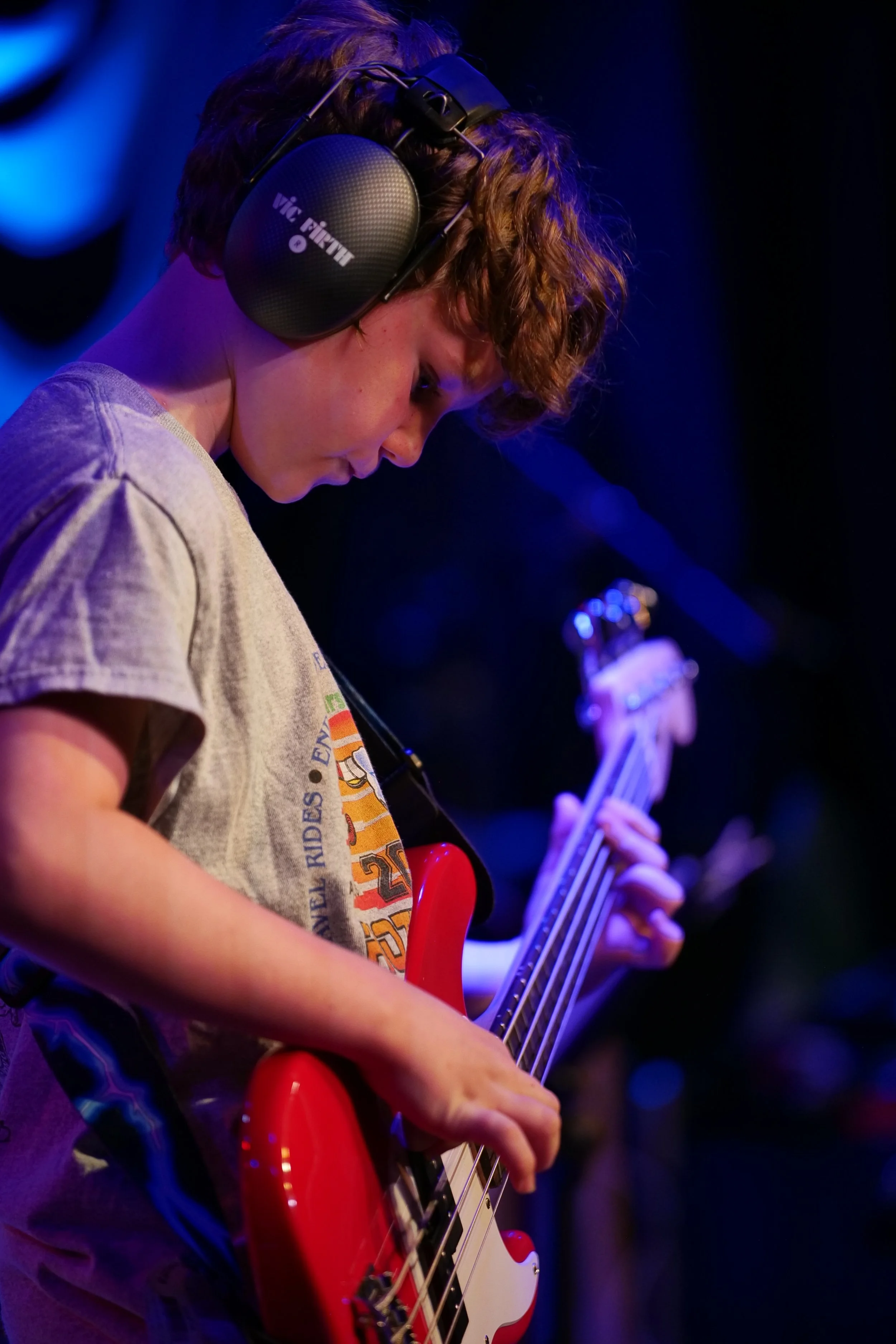 A young person with curly hair wearing headphones playing a red bass guitar on stage under blue lighting.