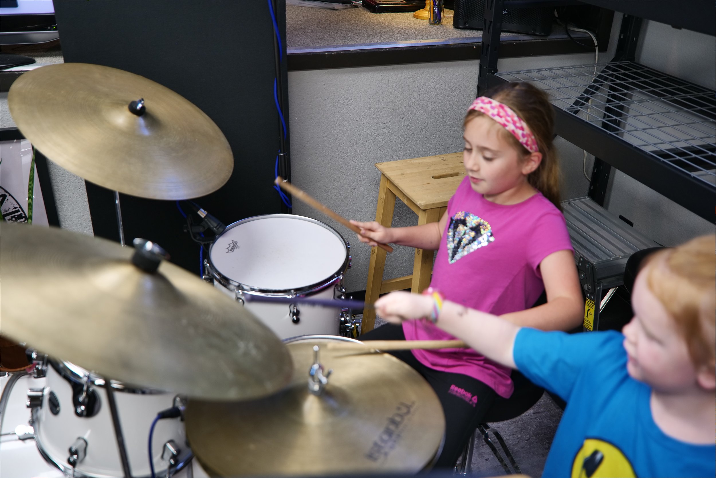 Two young children playing drums in a room, one girl wearing a pink shirt with a heart design and a pink headband, and one boy wearing a blue shirt, both holding drumsticks.