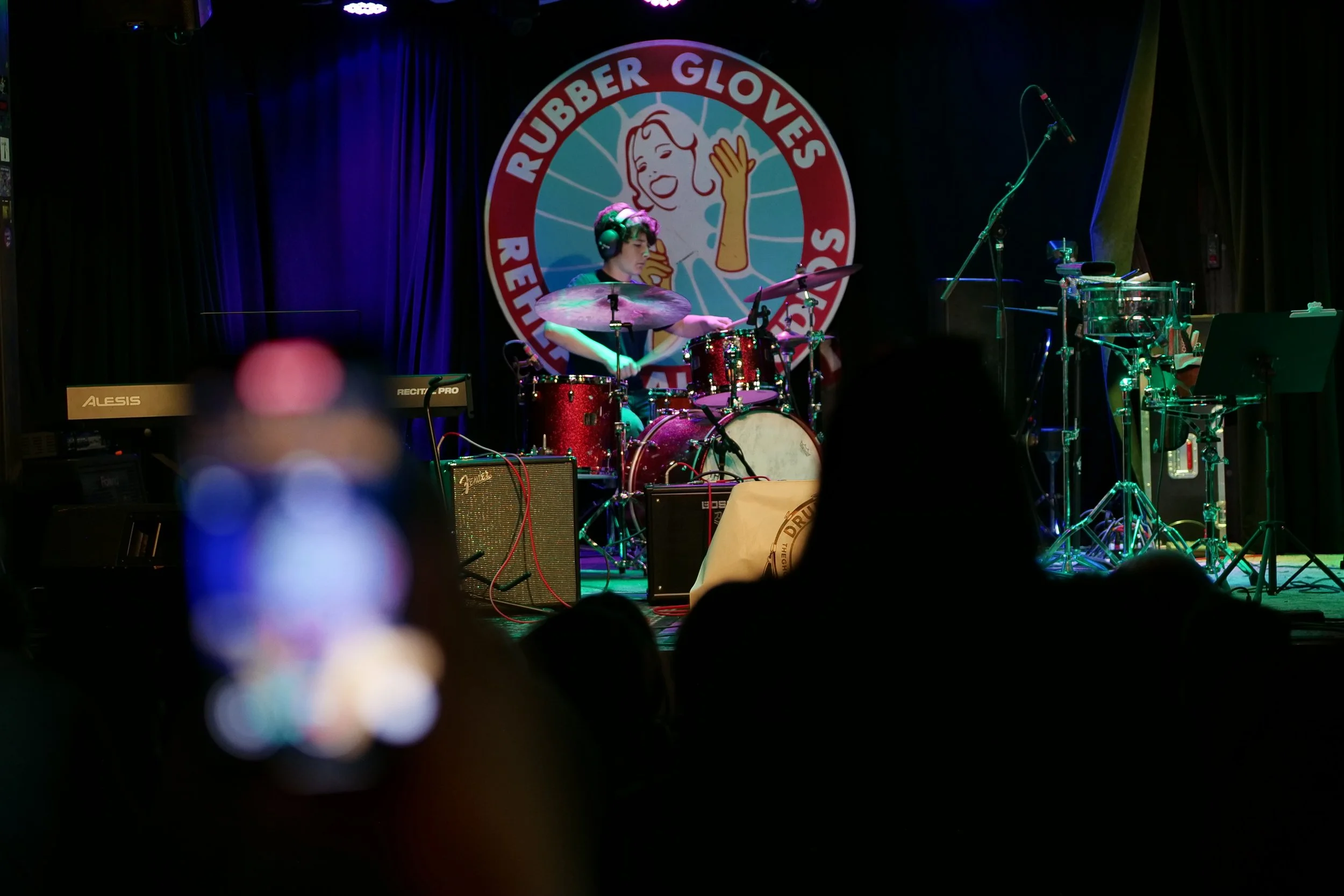 A musician playing drums on stage at Rubber Gloves Rehearsal Studios, with audience silhouette in foreground and a colorful logo in the background.