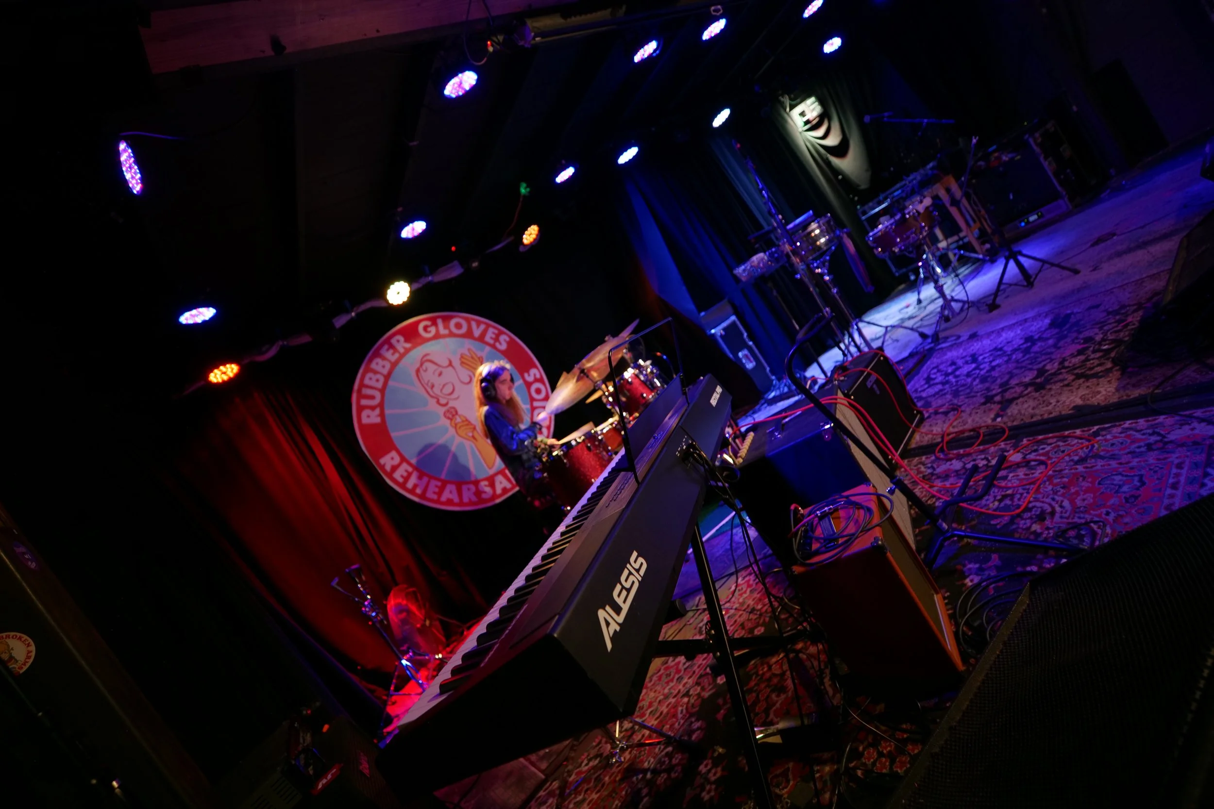 A musician playing drums on stage during a rehearsal, with a keyboard and other musical instruments around, in a venue with red curtains and stage lighting.