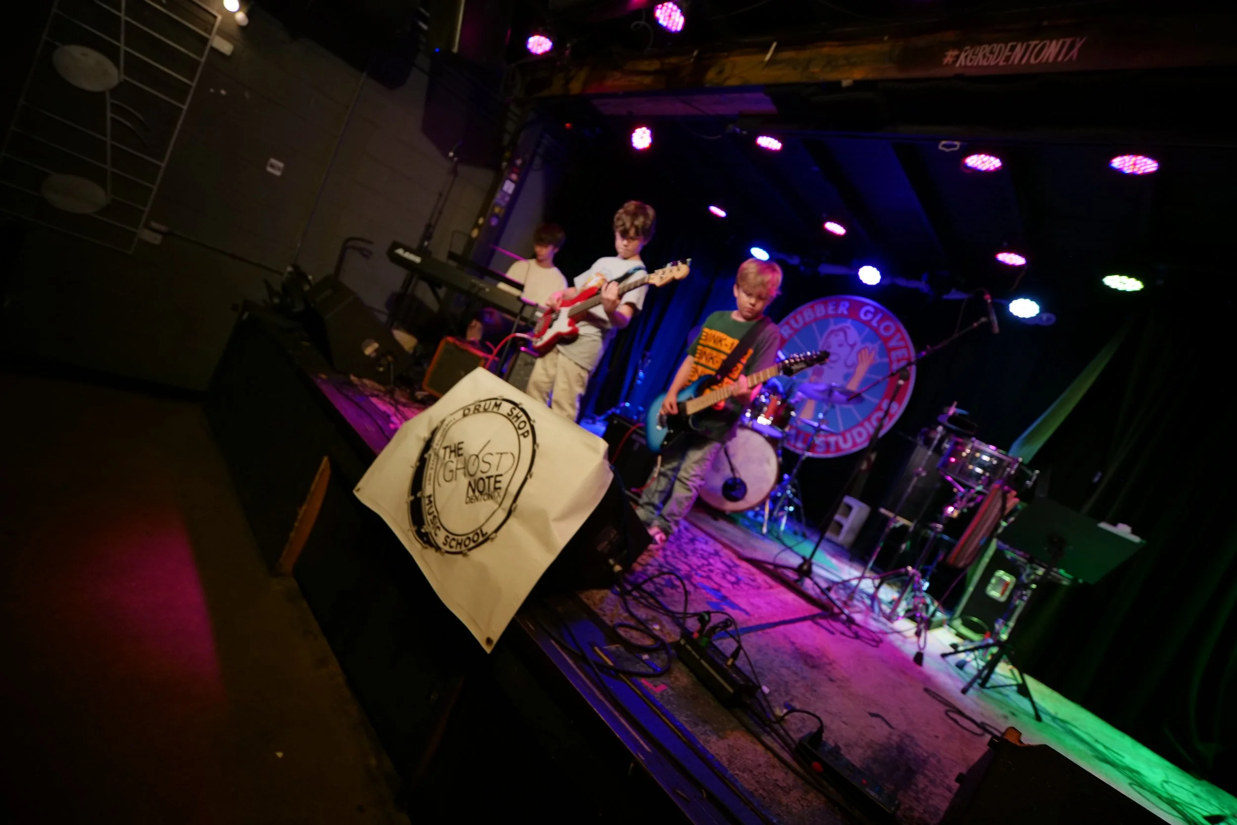 Young boys playing electric guitars and keyboard on stage during a concert at a music venue, with colorful stage lighting and music equipment visible.
