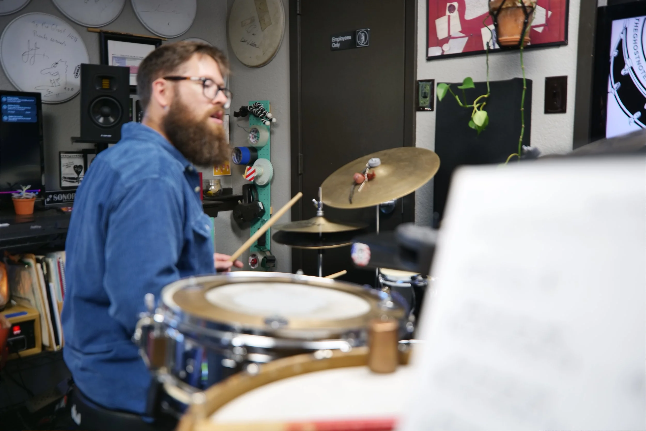 A man with glasses and a beard wearing a blue denim shirt playing drums in a music studio decorated with wall art, plants, and audio equipment.