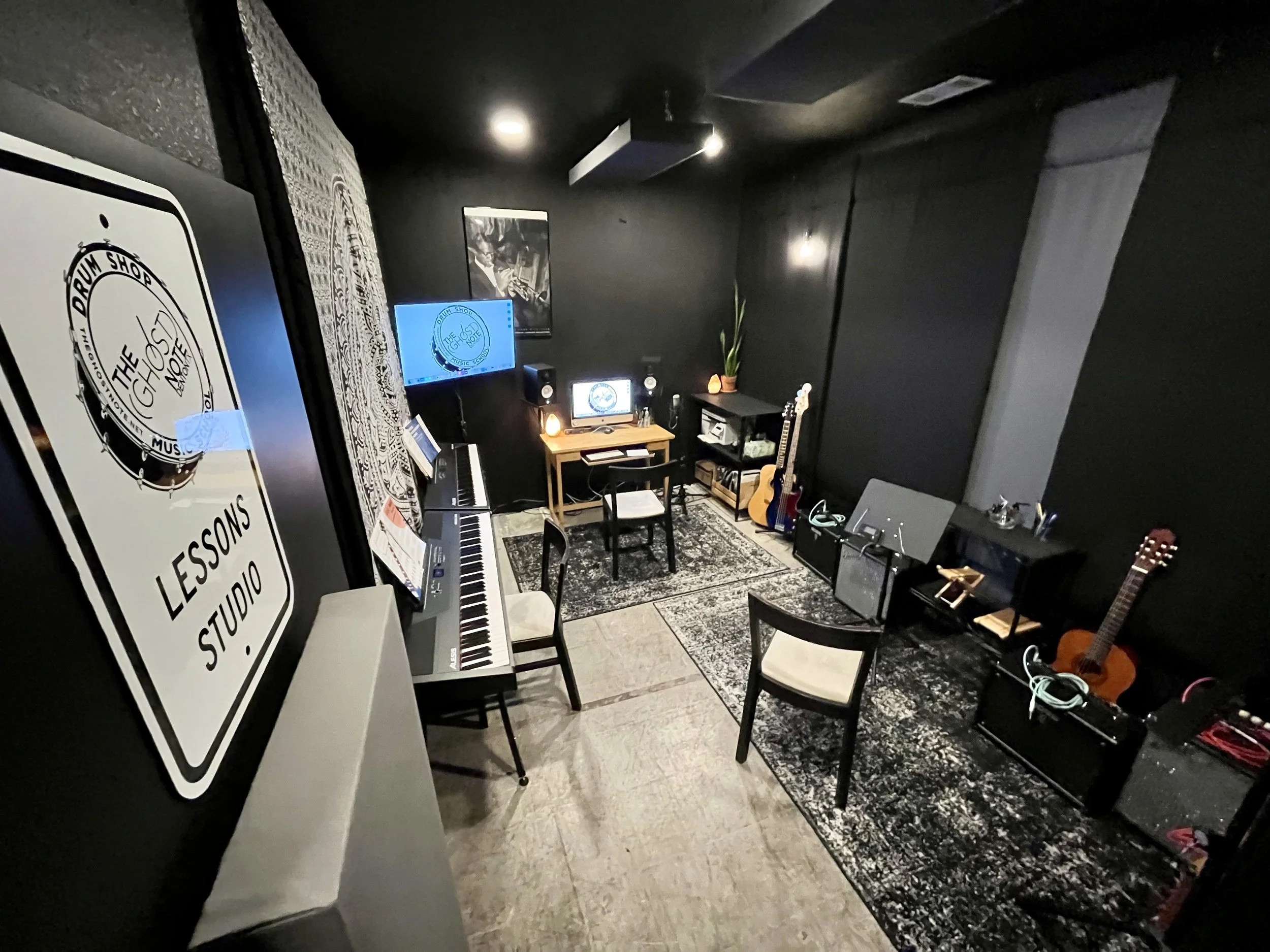 Music studio with black walls, a digital piano, guitars, a computer on a small desk, and various music equipment including speakers and amplifiers.