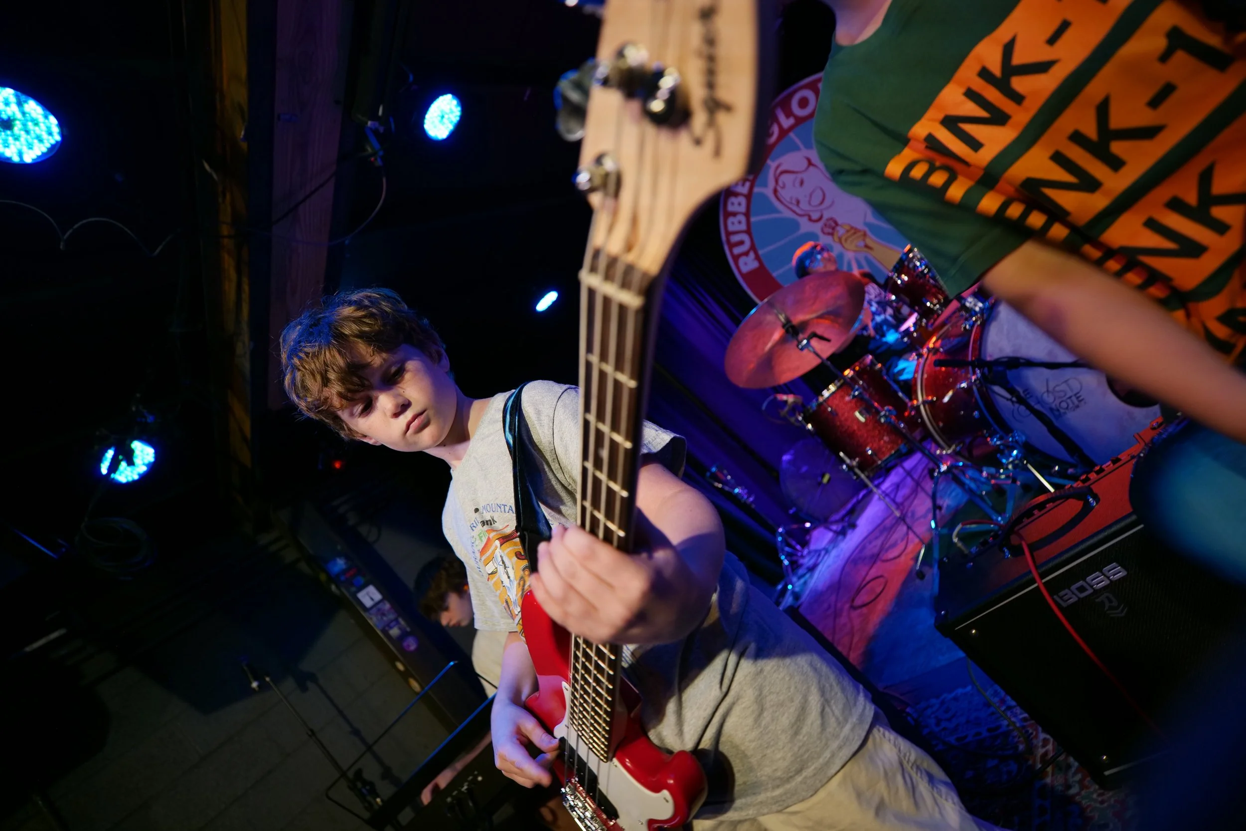 Young boy playing a red electric guitar on stage with drums behind him, stage lights overhead, in a dimly lit music venue.