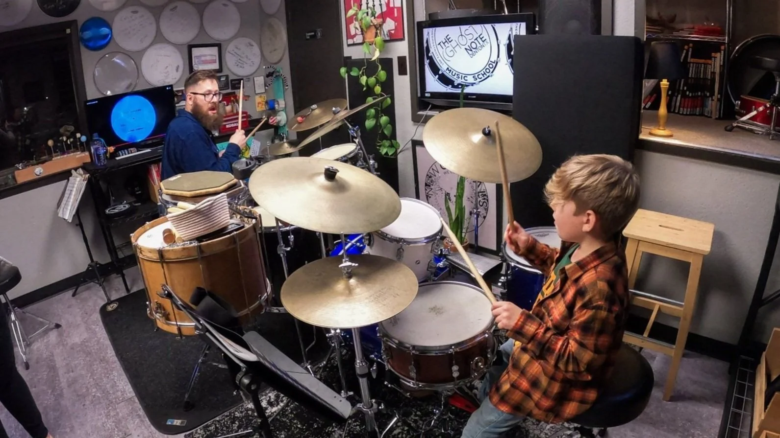 A young boy practicing drums in a music classroom with a man in the background, both seated at drum sets, surrounded by musical equipment and posters.