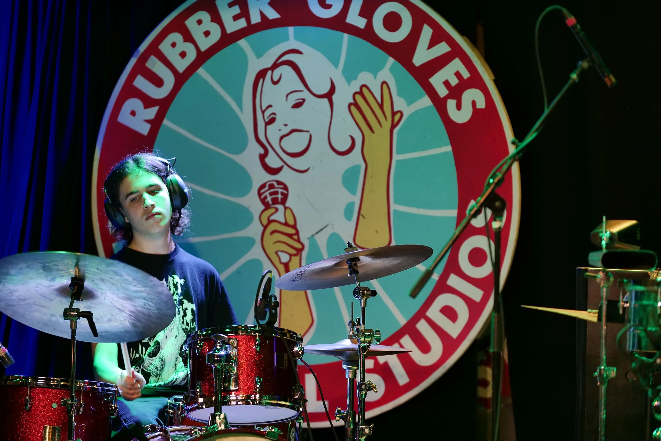 A young man with headphones playing the drums on stage, with a large circular logo in the background that says 'Rubber Gloves' and features a cartoon woman holding a microphone.