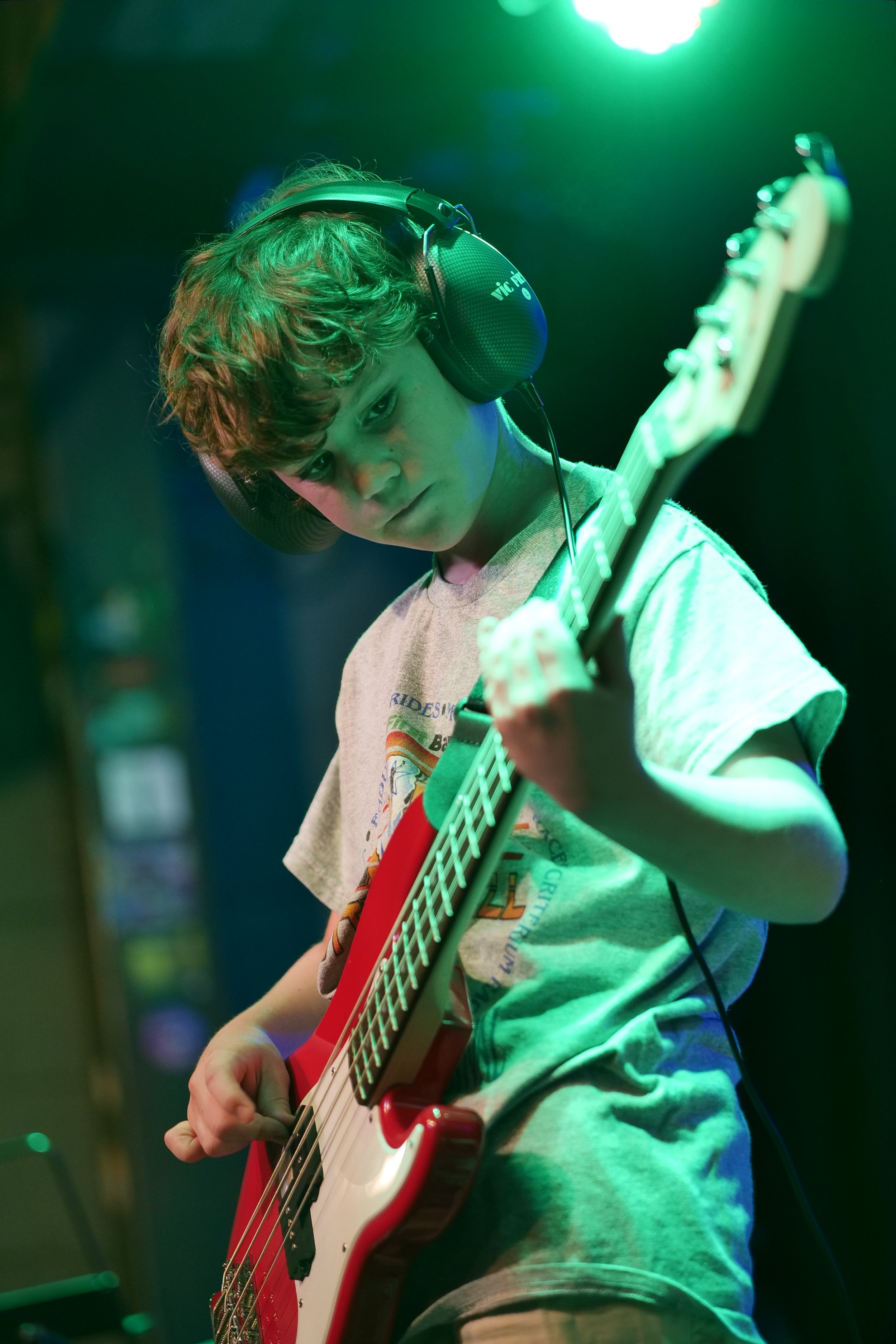 A young boy with curly hair wearing headphones, playing a red and white electric bass guitar, with a focused expression, in a setting with green and blue lighting.