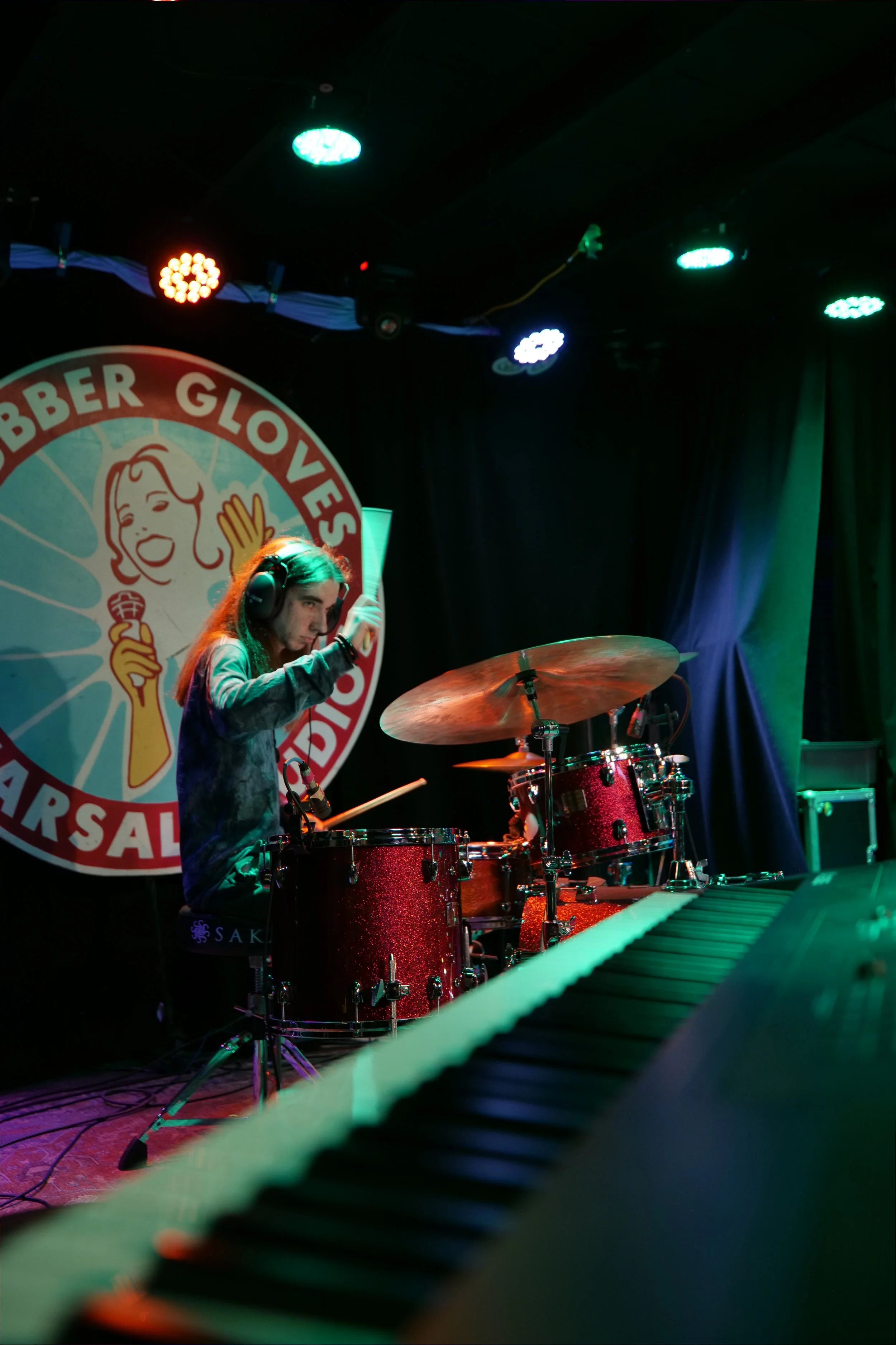 A young woman playing drums on a stage with neon lights. The background features a large circular logo with a smiling woman holding an ice cream cone and the words "Glover's Marsala". The scene is dimly lit with colorful stage lights.