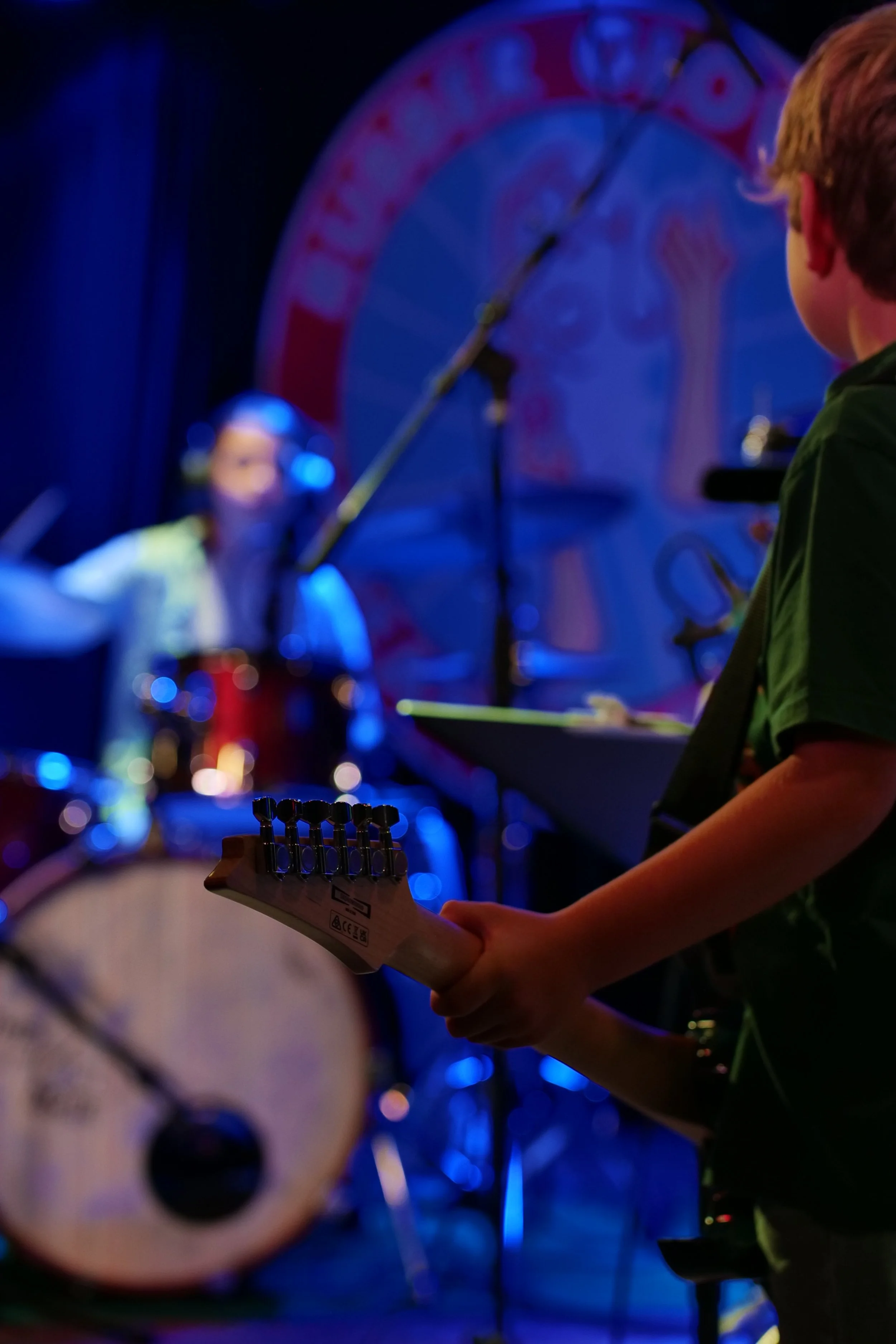 A young musician playing an electric guitar on stage with drums and a colorful backdrop in the background.
