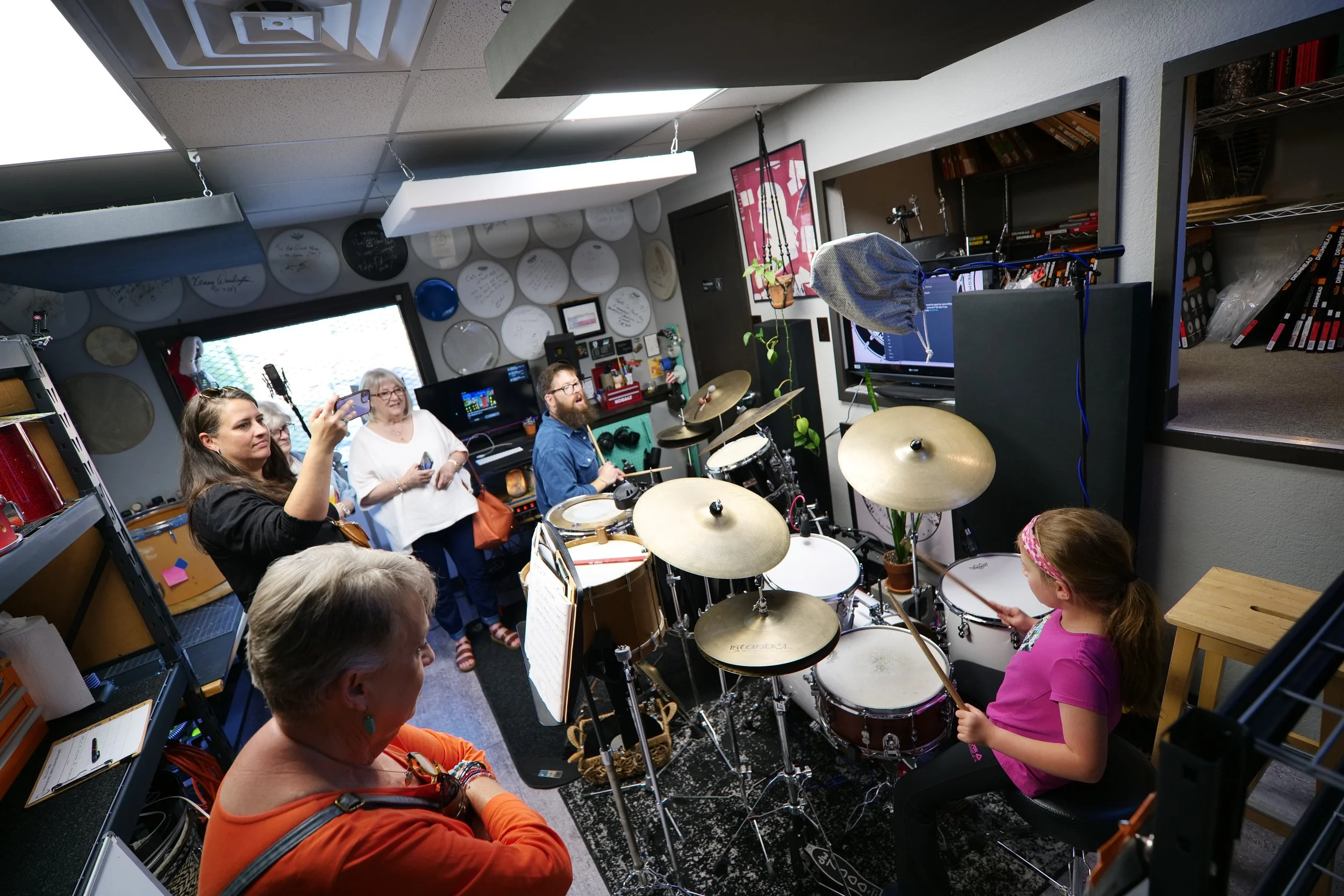 A group of people in a music studio gathered around a girl playing the drums, with some taking photos and others watching, surrounded by musical instruments and equipment.