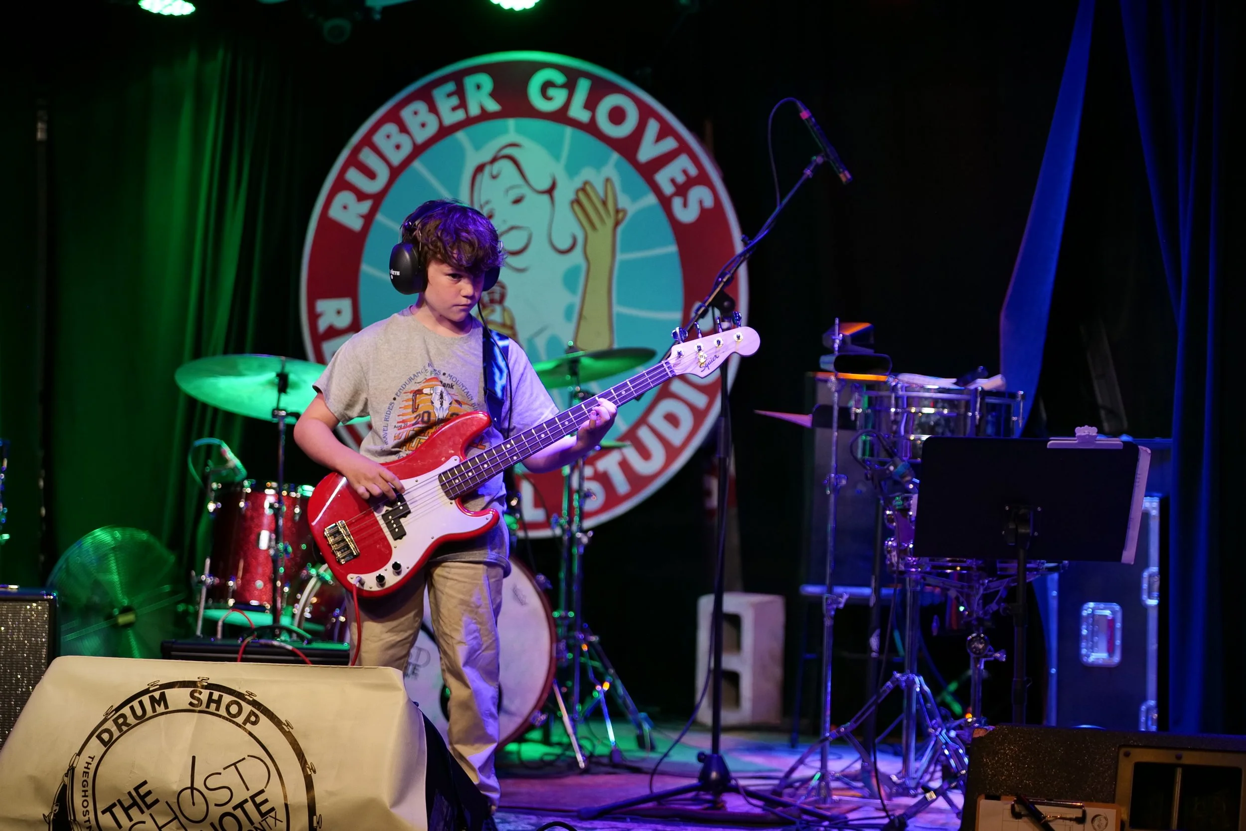 A young boy playing a bass guitar on a stage with musical equipment, microphones, and a large logo behind him that reads 'Rubber Gloves Studio.'