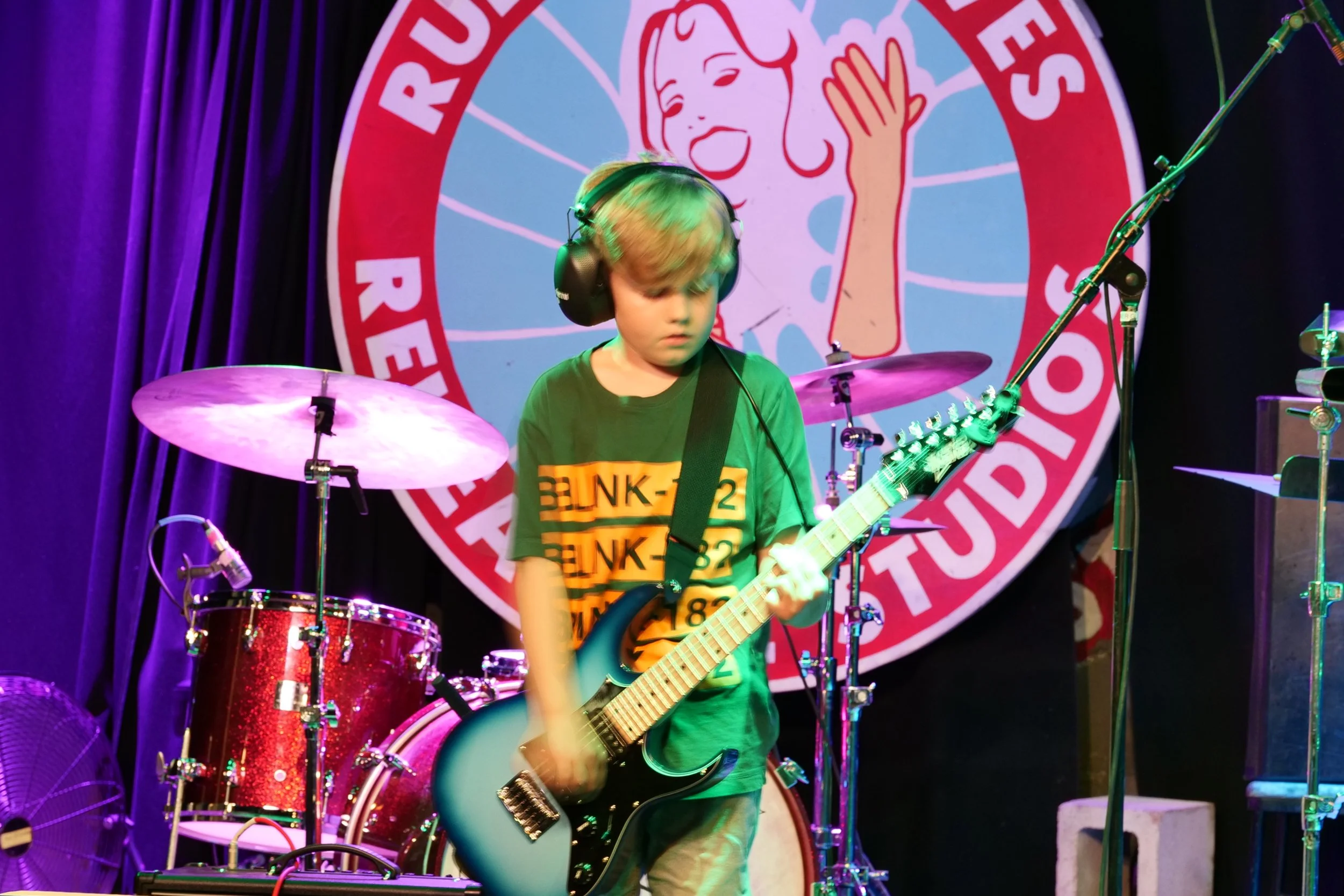 Young boy playing an electric guitar on stage with drums behind him and a large circular sign with a cartoon character in the background.