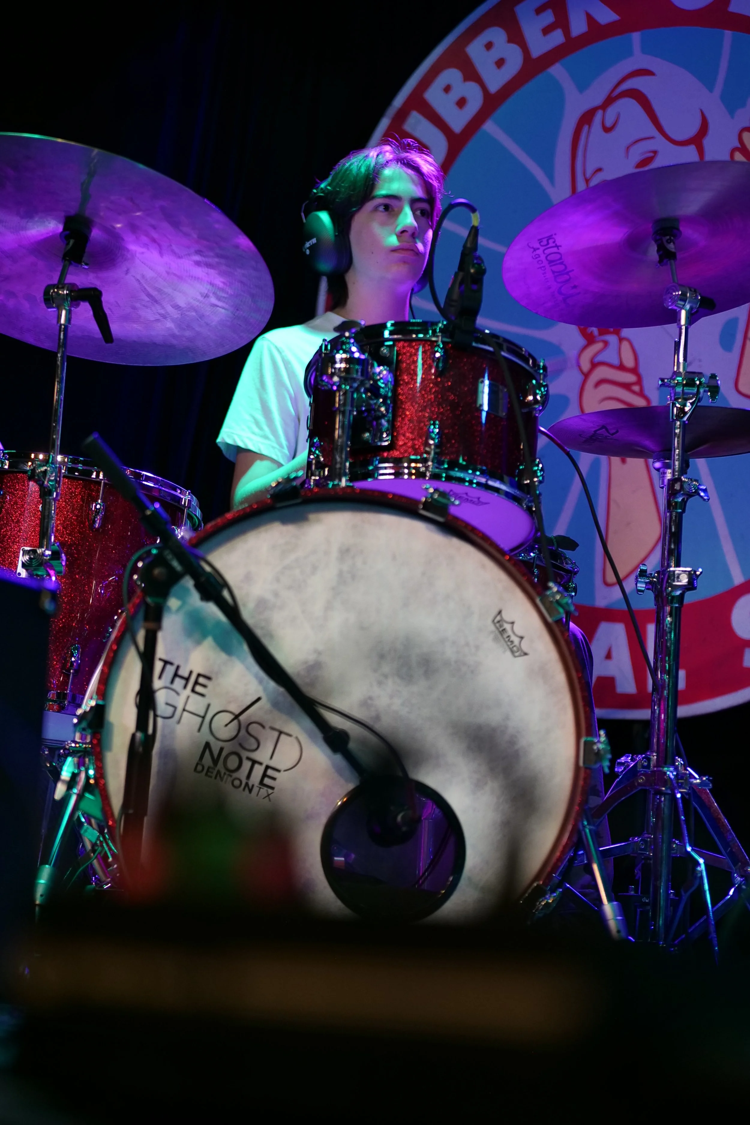 A young musician with headphones playing a red drum set on stage, with a backdrop featuring a logo with cartoonish artwork.