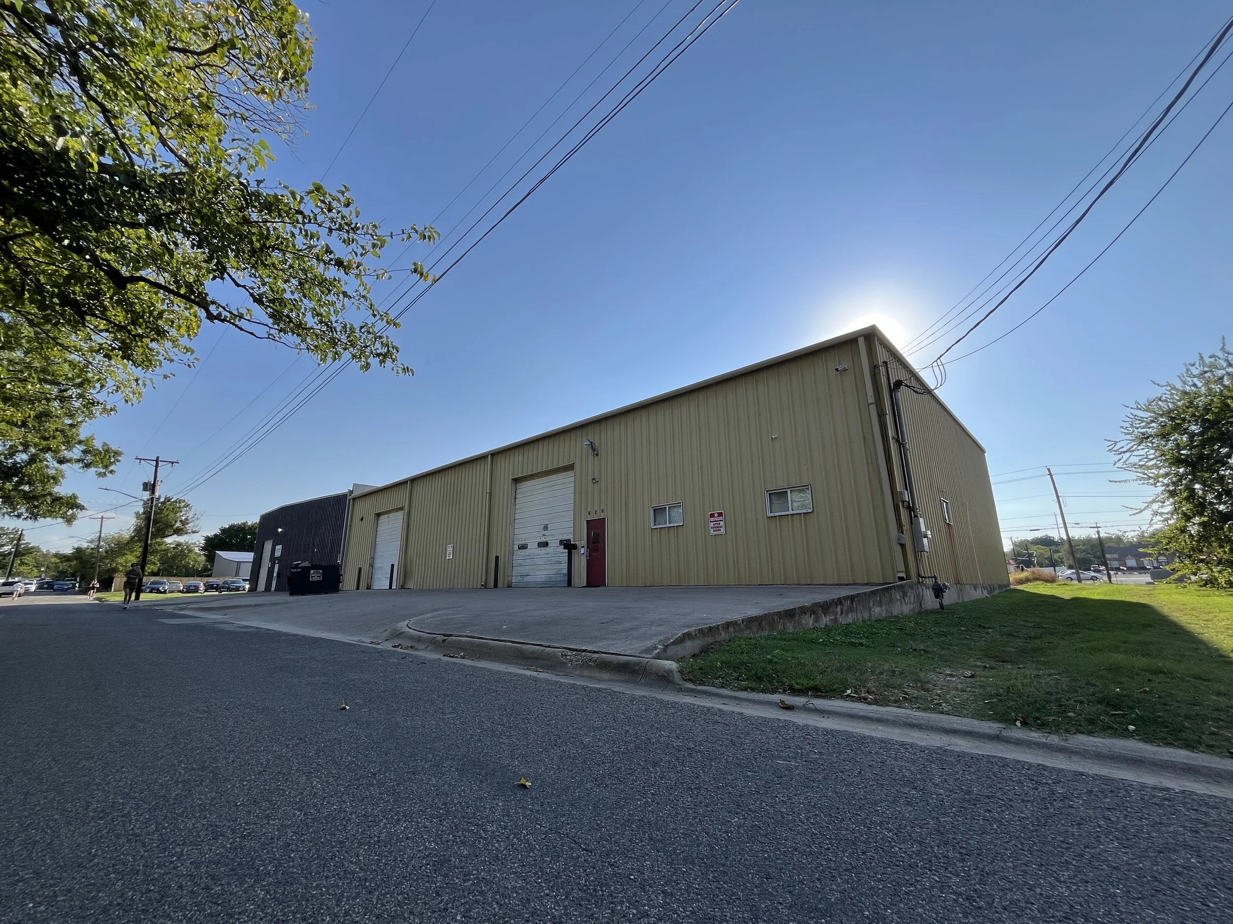 A beige industrial building with two large garage doors and a small red door, situated on a slight hill with a concrete driveway. There is a green grassy area to the right, and a man walking on the street. Overhead power lines and trees are visible a