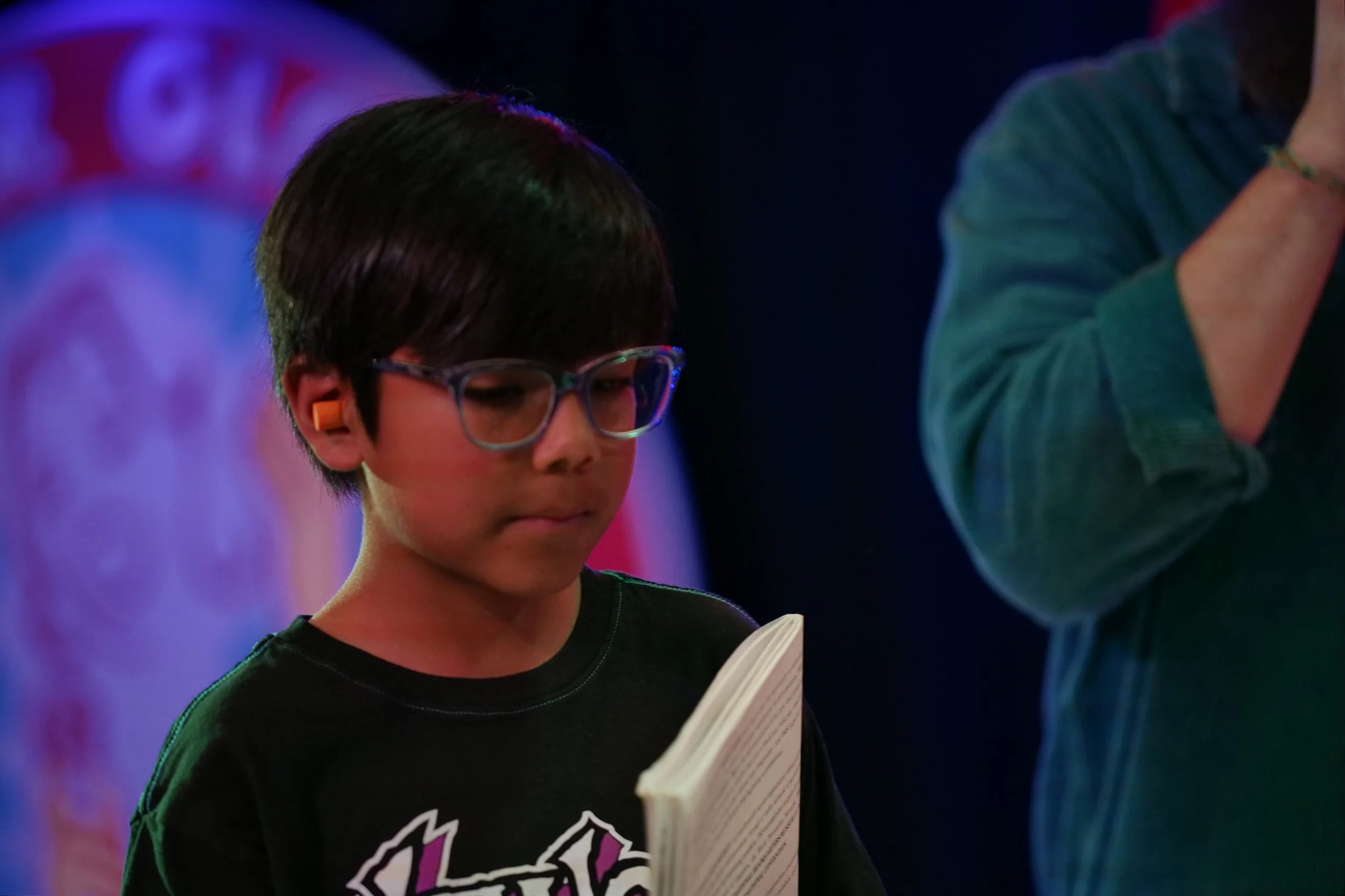 A young boy with glasses and earplugs reading a book in a dimly lit environment with colorful background lights.