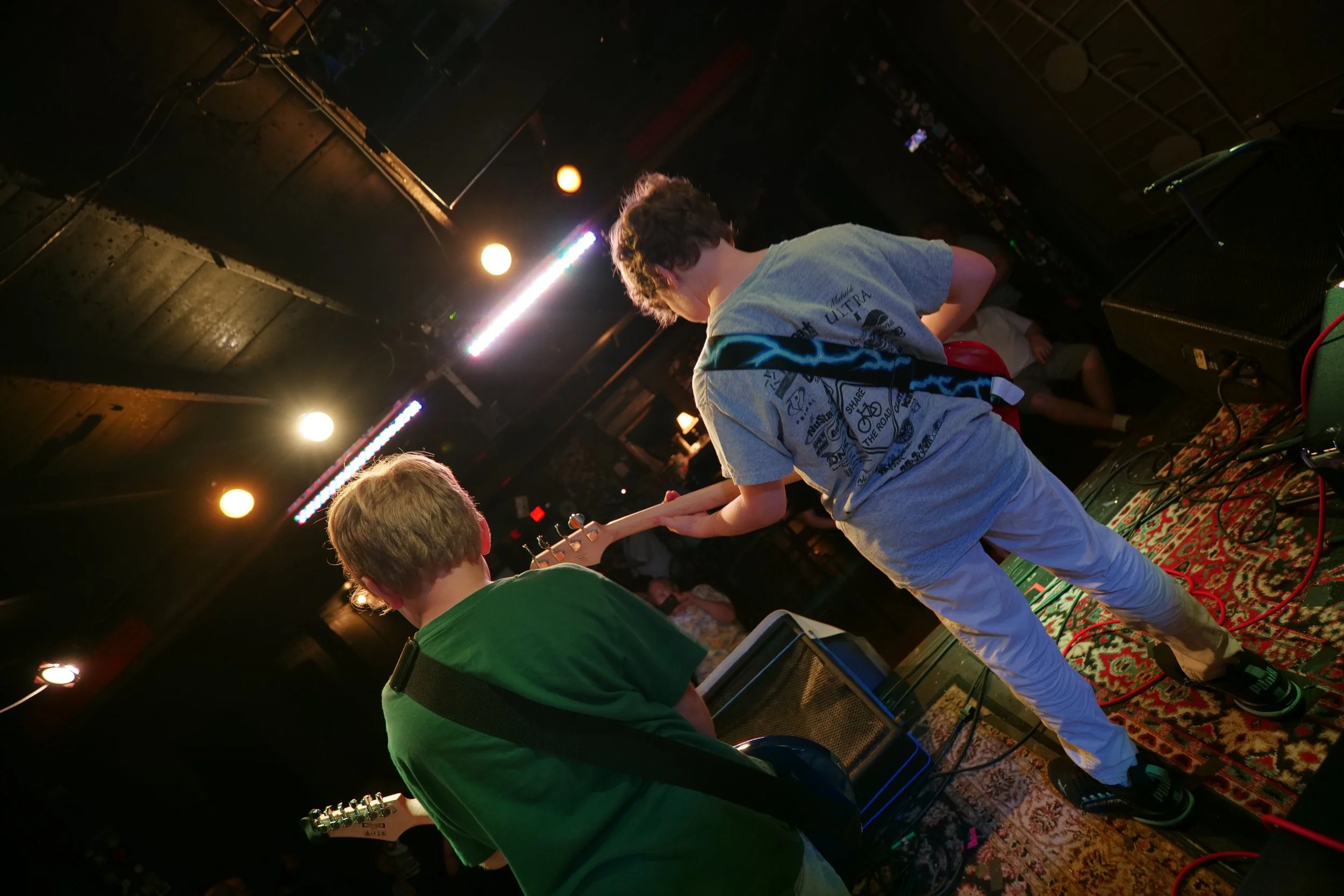 Two young boys playing electric guitars on a stage with colorful stage lights overhead. The scene captures a casual music performance in a dimly lit venue.