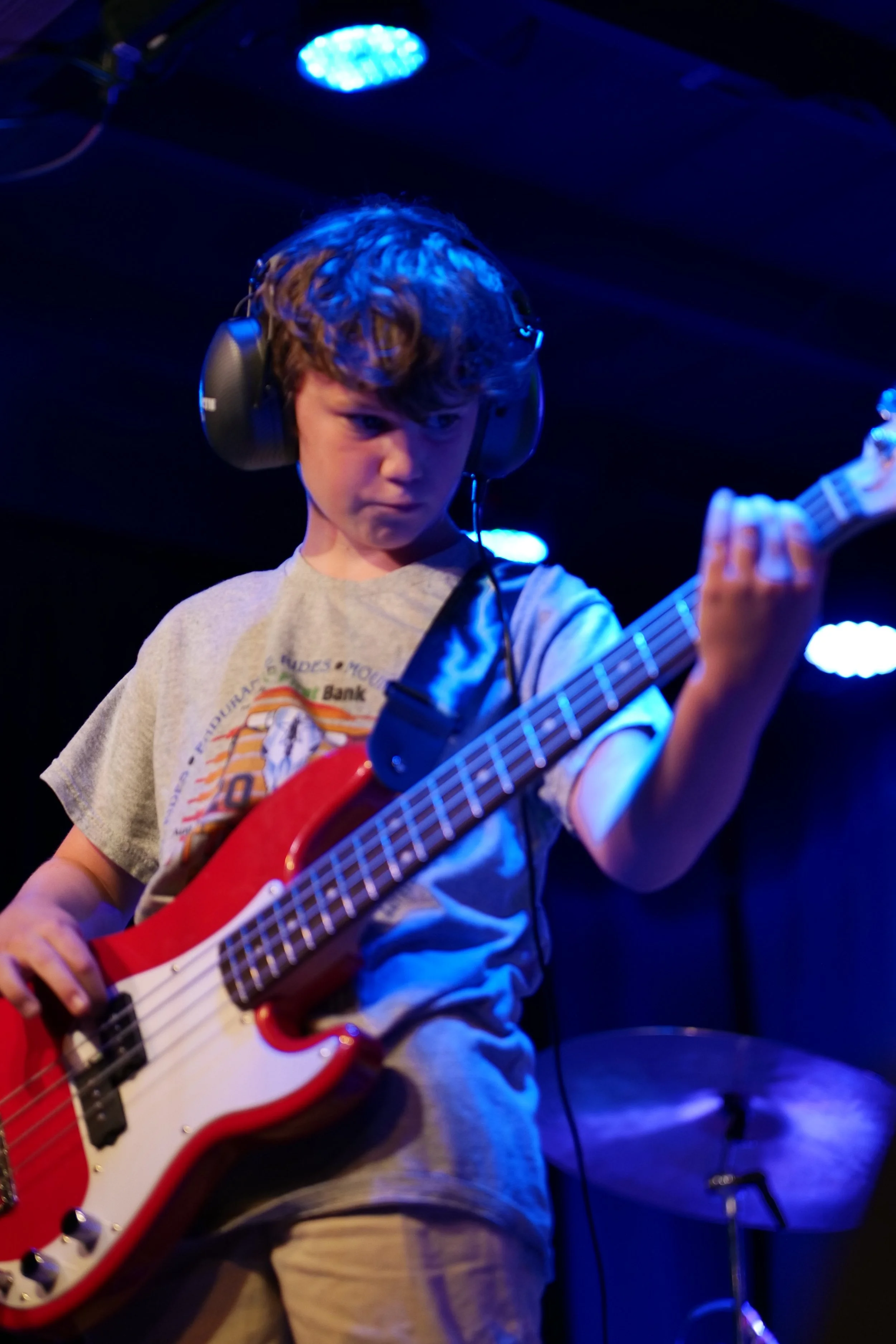 Young boy wearing headphones playing a red electric bass guitar on stage with blue lighting.