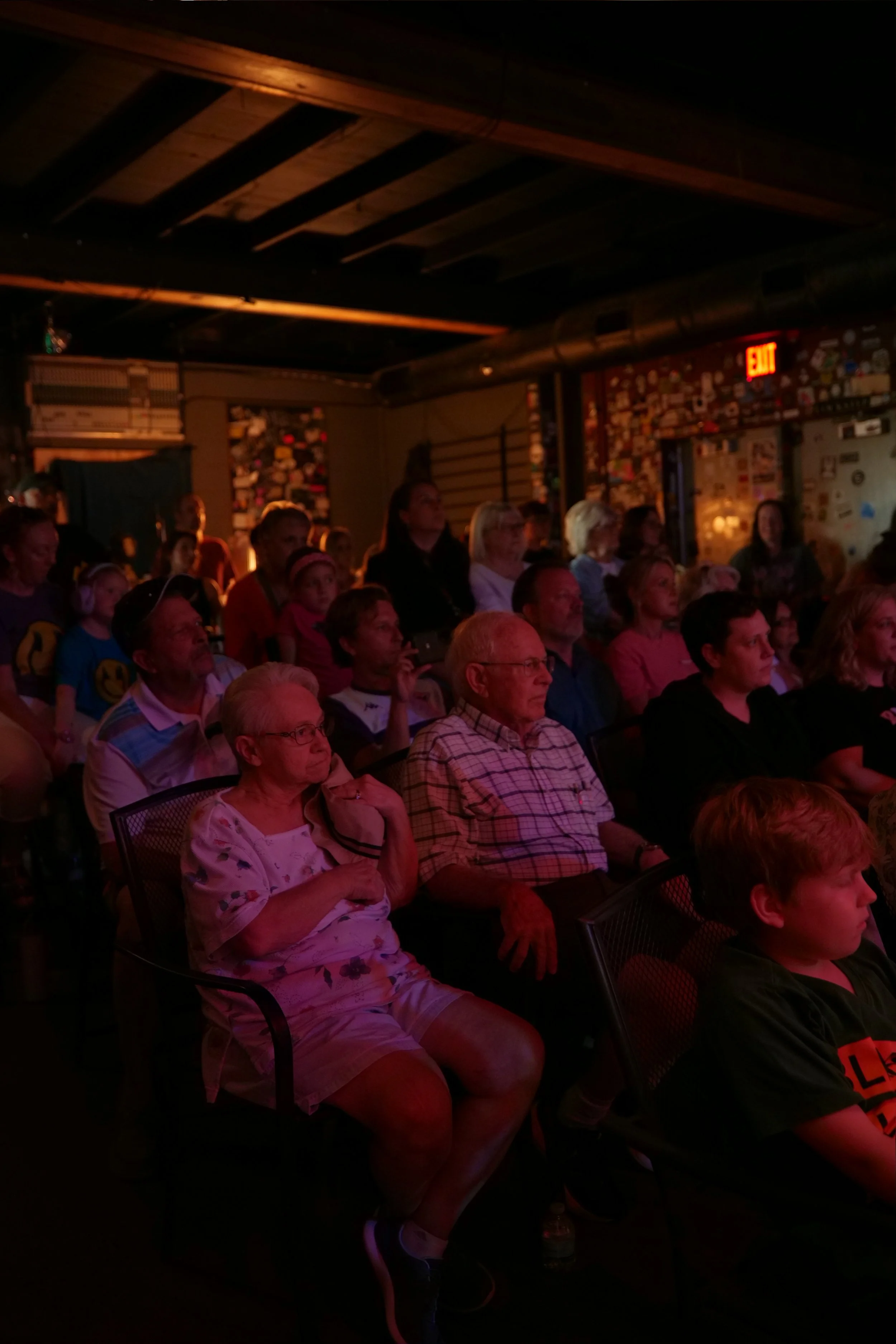 Audience sitting in a darkened room watching a performance or presentation, with a wall decorated with various stickers and an illuminated exit sign in the background.