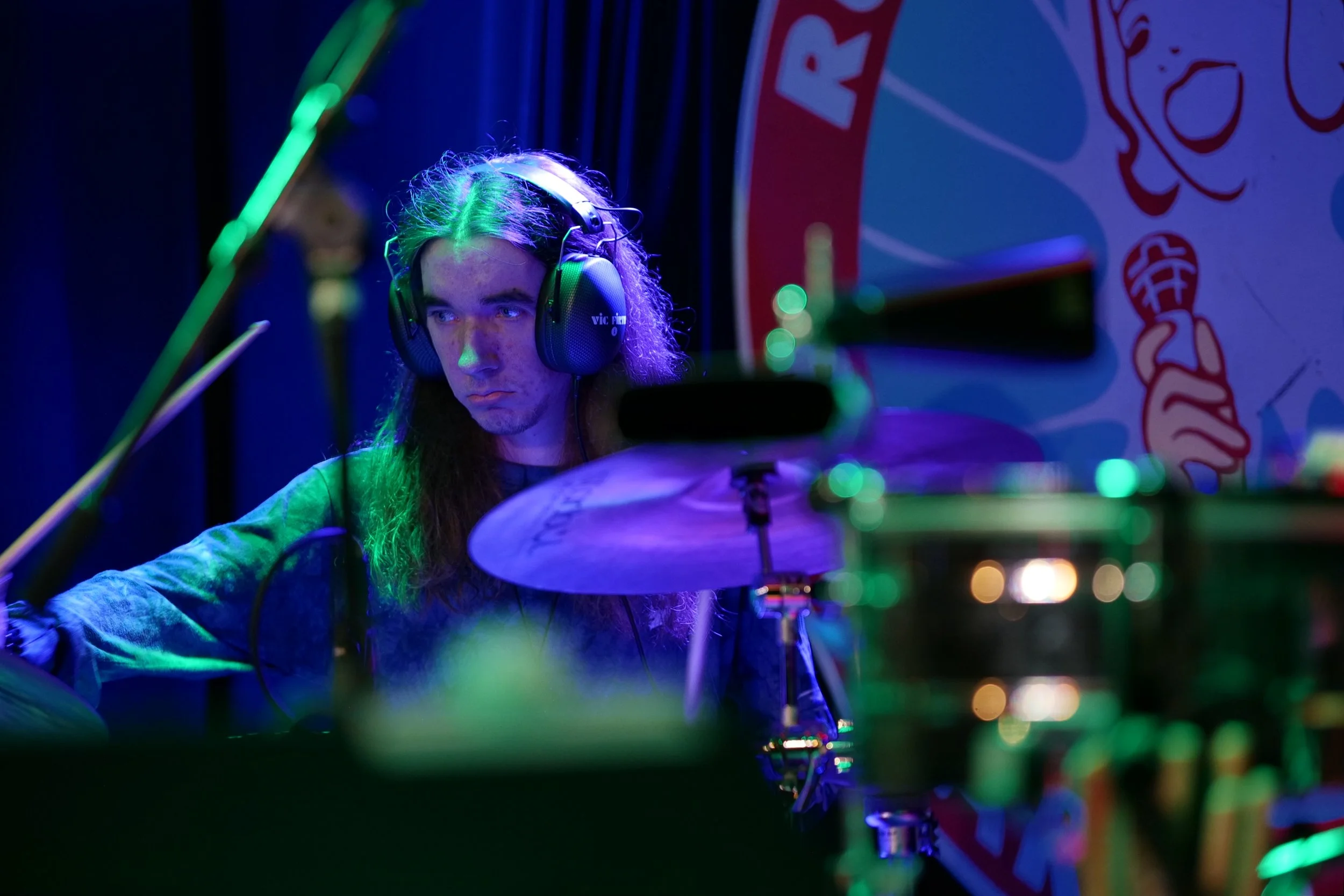 A young man with long hair wearing headphones while playing drums on stage, illuminated by blue-green lighting, with a colorful backdrop featuring graphics and text.