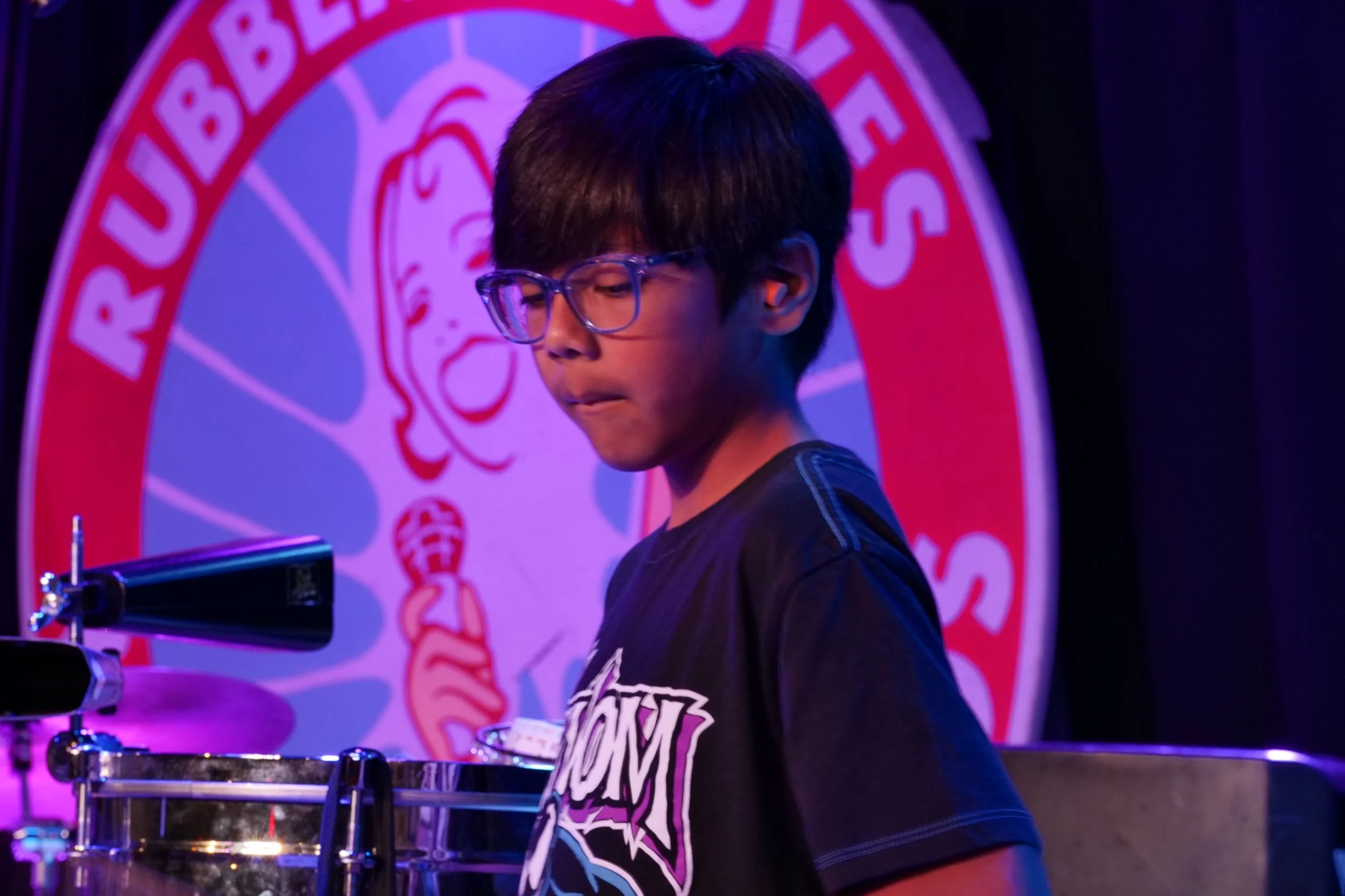 A young boy with glasses playing percussion on stage at a comedy club, with a large circular logo of a cartoon girl holding a microphone in the background.
