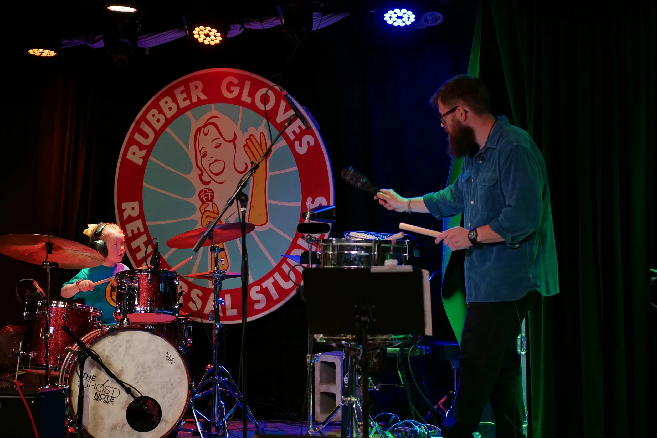 A young girl playing drums on stage with a man operating electronic music equipment. The stage features a large circular logo for Ruben Glovers Rebel Studio in the background with a smiling woman and a hand gesture.