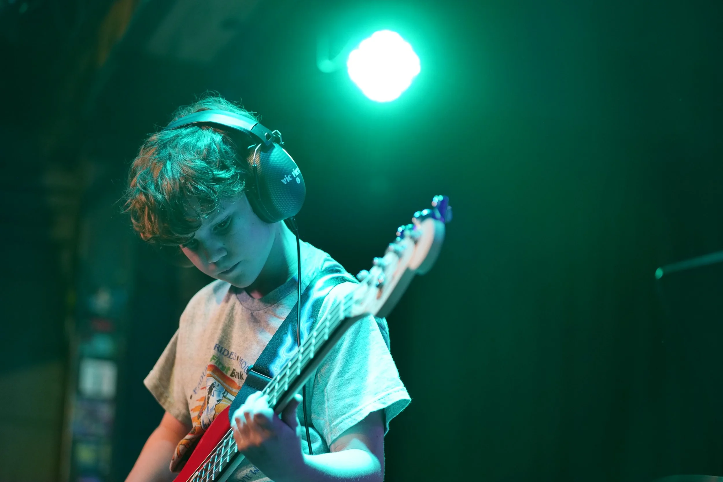 A young boy with curly hair wearing headphones, playing an electric guitar on stage with green and blue lighting.