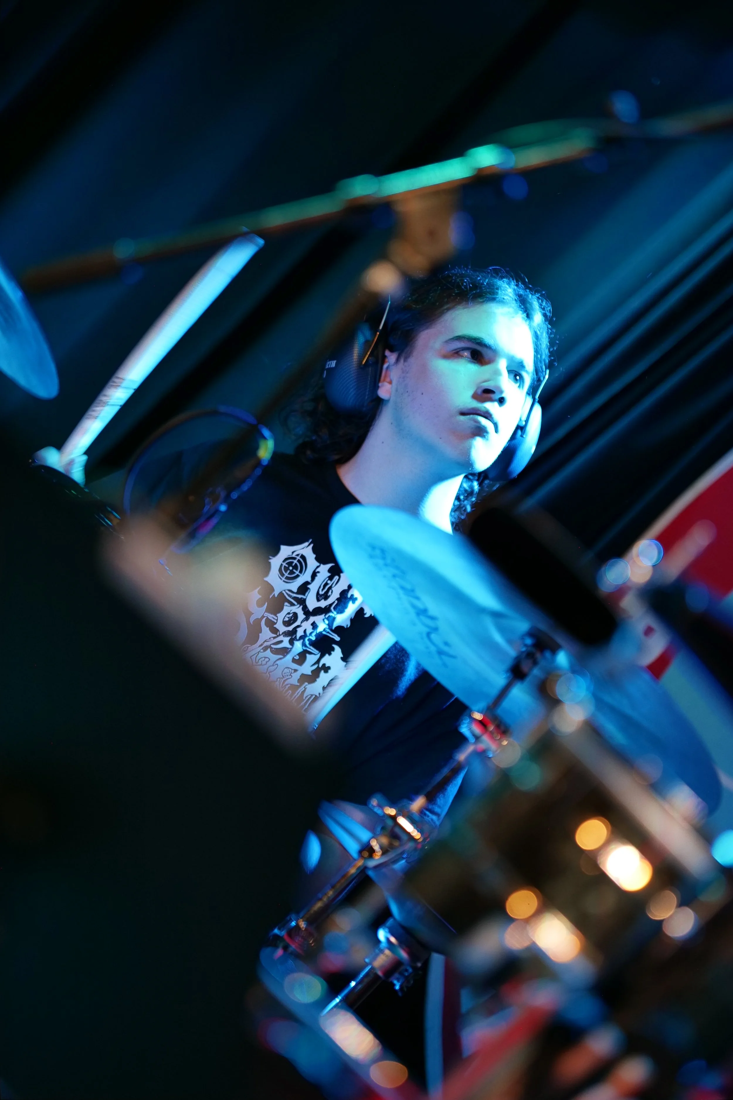Young musician playing drums, wearing headphones, with a focused expression, in a dimly lit setting with blue lighting.