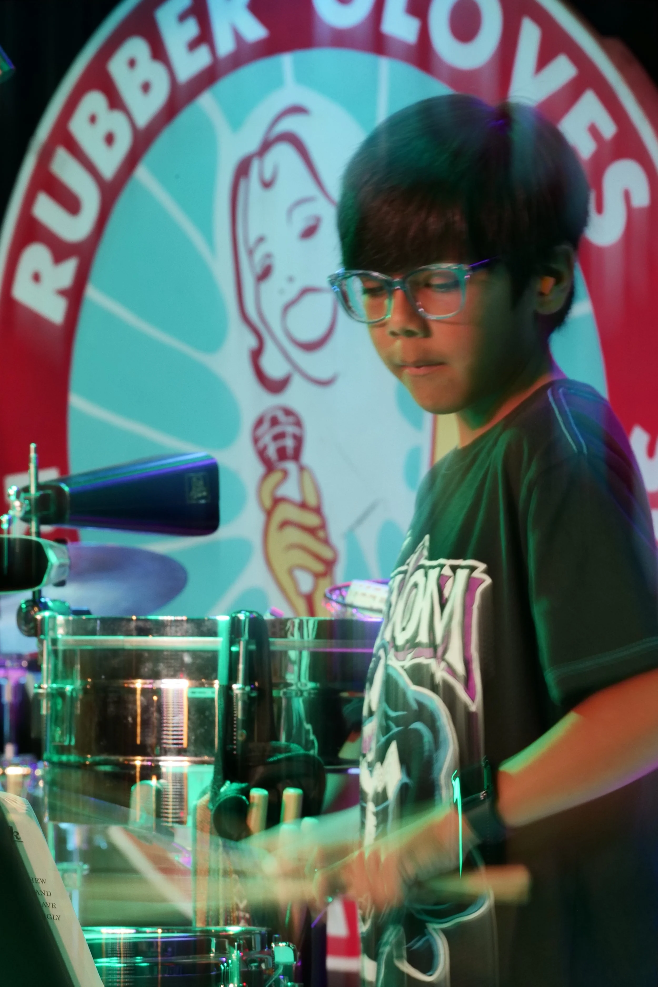 A young man with glasses playing a drum set at a musical event, with a colorful logo featuring a woman holding a microphone in the background.
