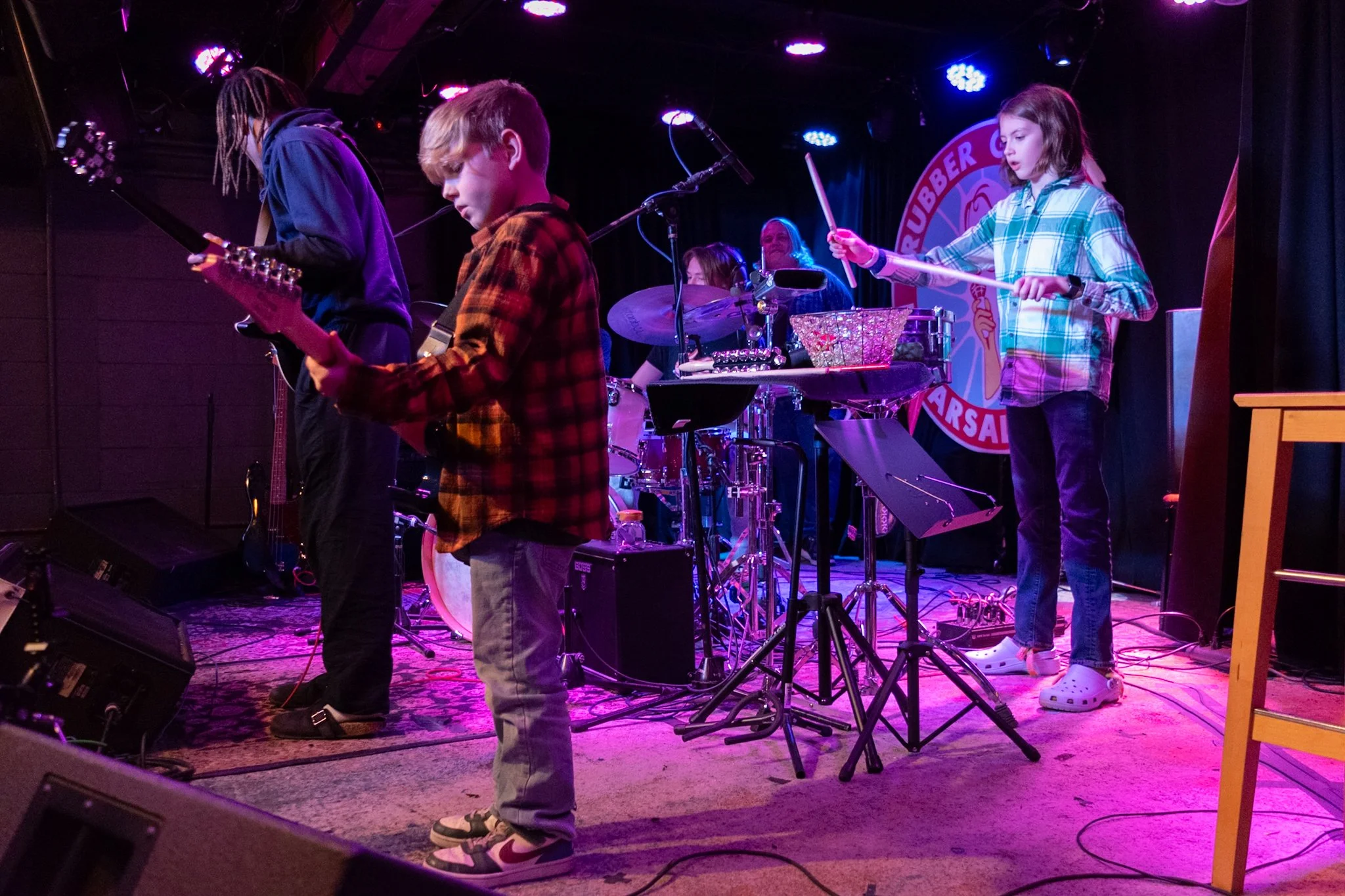 Children performing on stage with musical instruments, colorful stage lighting, and a sign in the background that reads 'Rubber City Dar Salvation.'